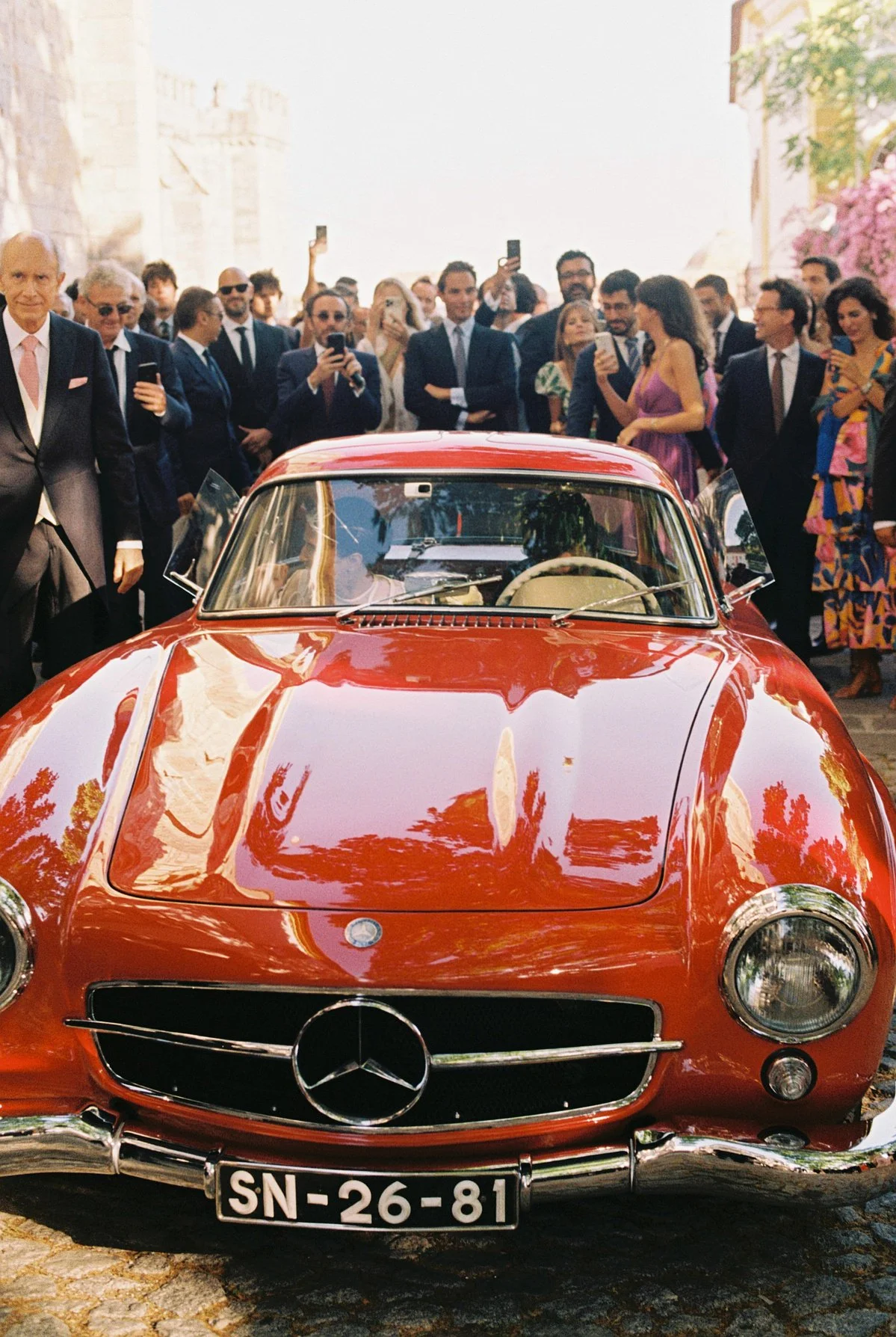 A shiny red vintage Mercedes-Benz sports car parked with a crowd of people gathered around, some taking photos, on a cobblestone street during daytime.