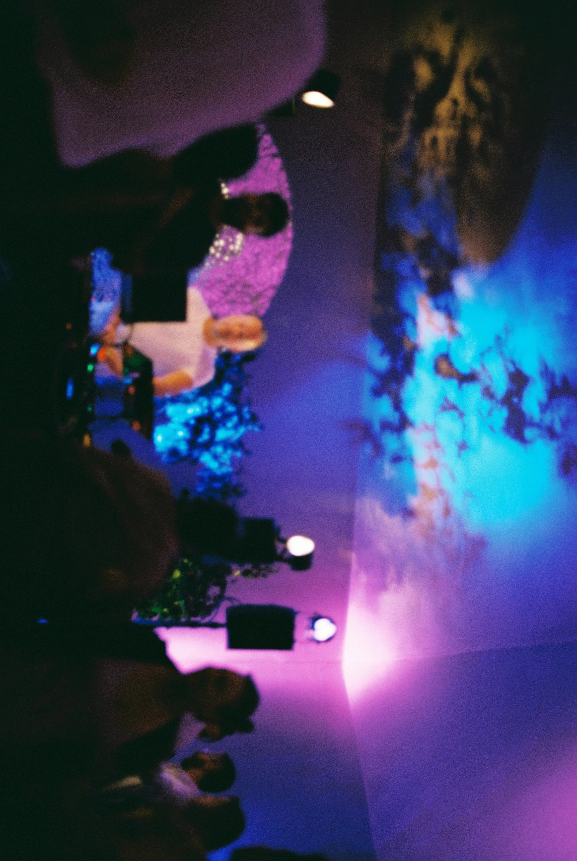 People watching a colorful outdoor movie or presentation at dusk, with purple, pink, and blue sky, and surrounded by trees and some glow lights.