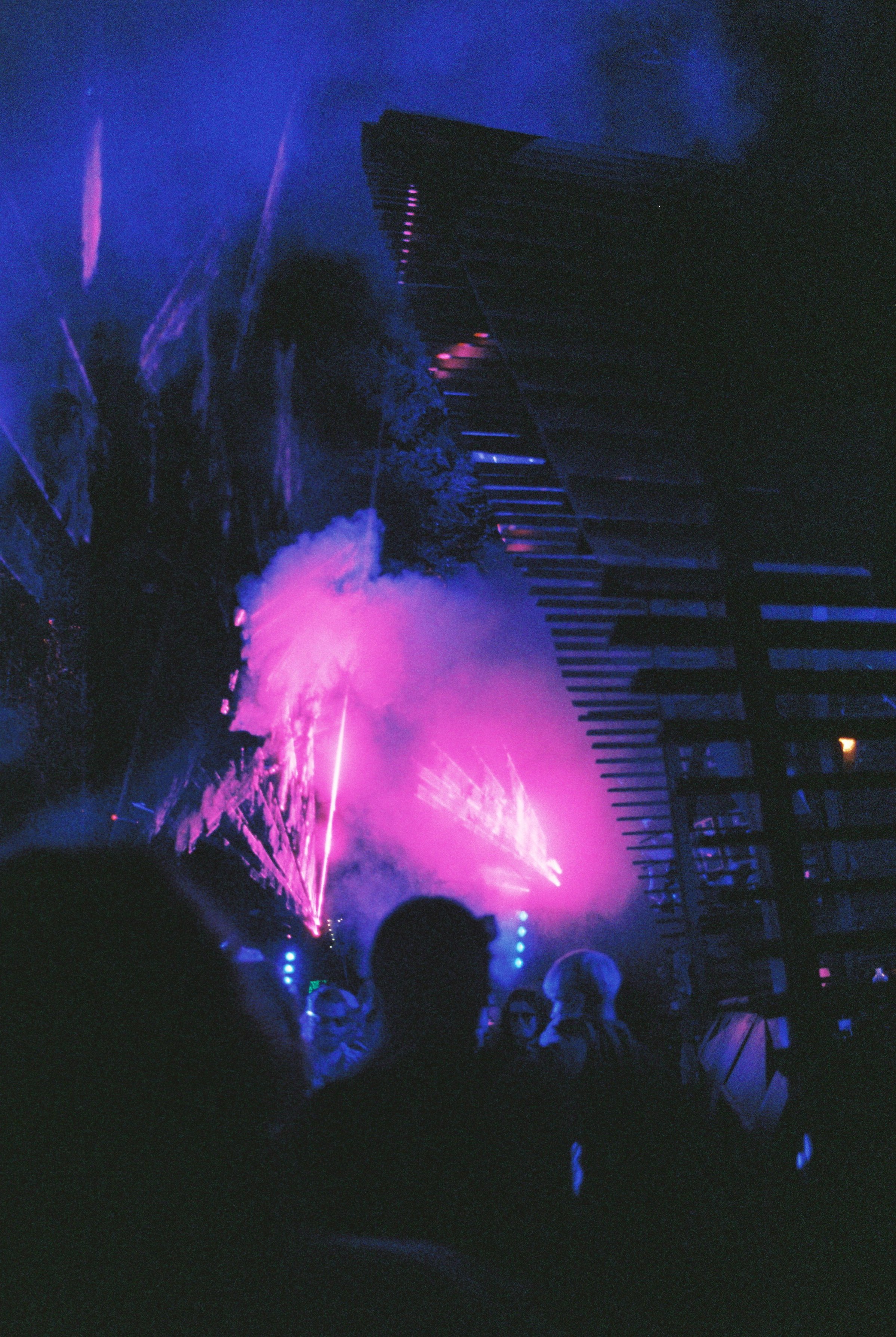 People watching a fireworks display at night with vibrant purple and blue lights illuminating the sky, some silhouettes visible in the foreground.