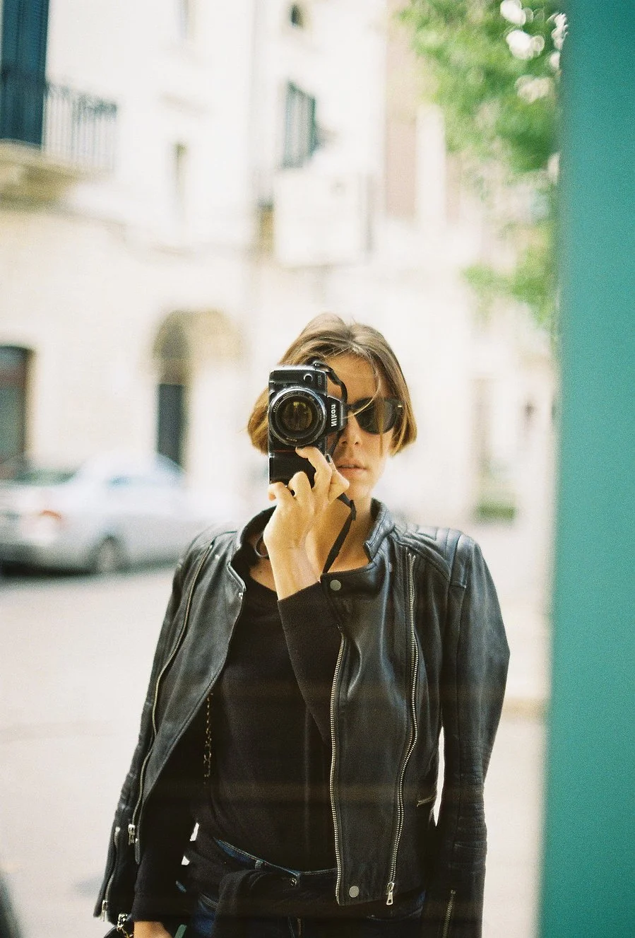 A woman taking a photo in a mirror, wearing sunglasses and a black leather jacket, with a blurred city street background.