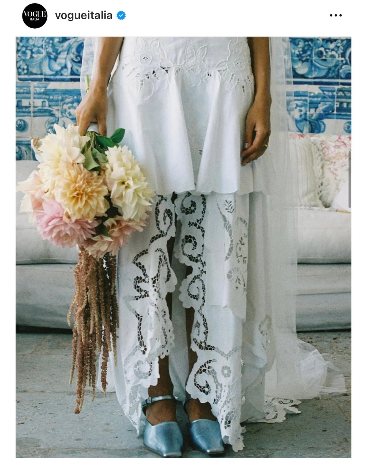 Lower half of woman in white embroidered dress holding a bouquet of flowers, standing on a stone surface near a light-colored sofa with floral pillows, against a backdrop with blue and white tile artwork.