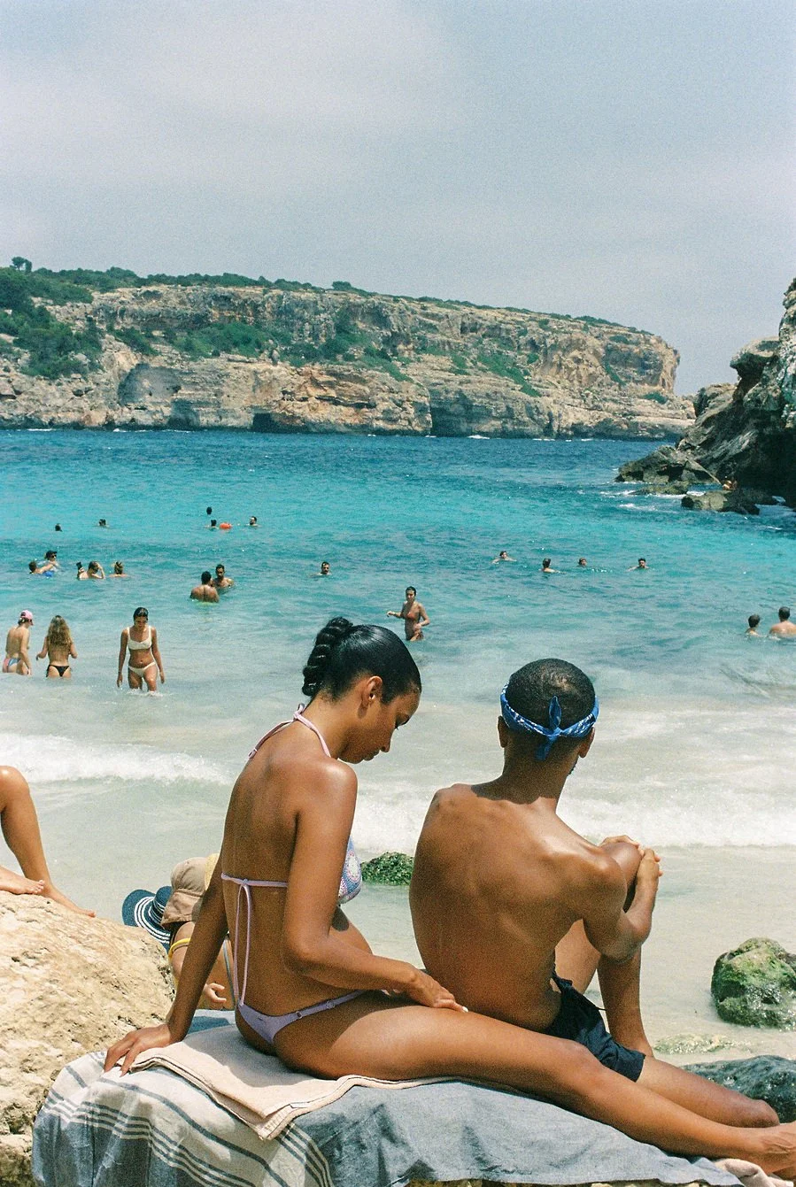 People relaxing on rocks and swimming in a beach with cliffs in the background.