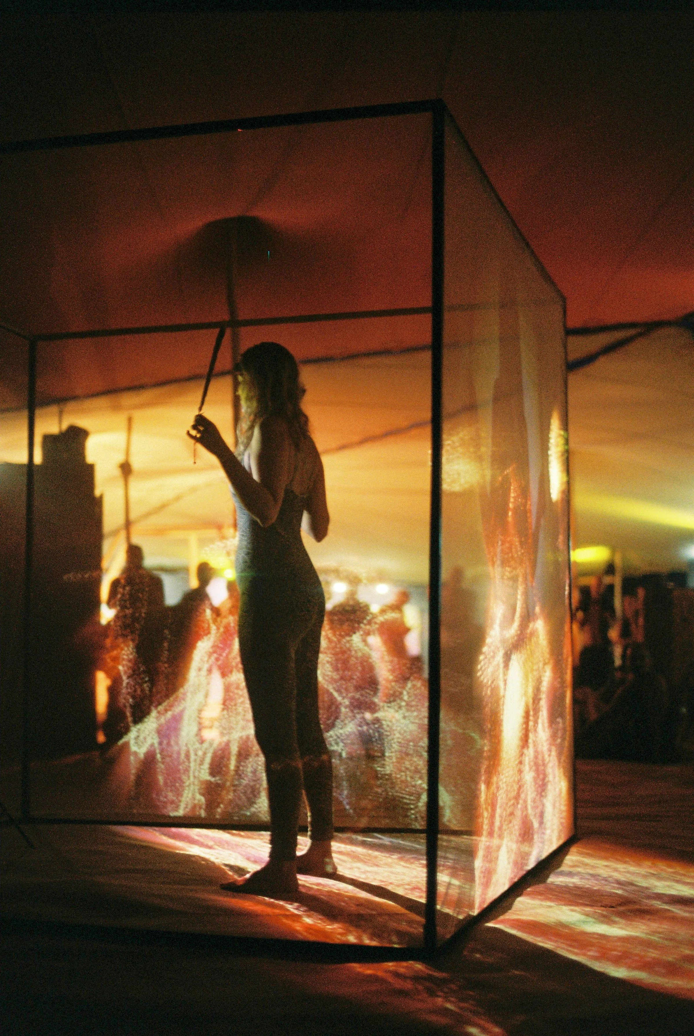 A woman standing inside a transparent cube, holding an umbrella, with warm lighting and blurred background, possibly at an event or exhibition.