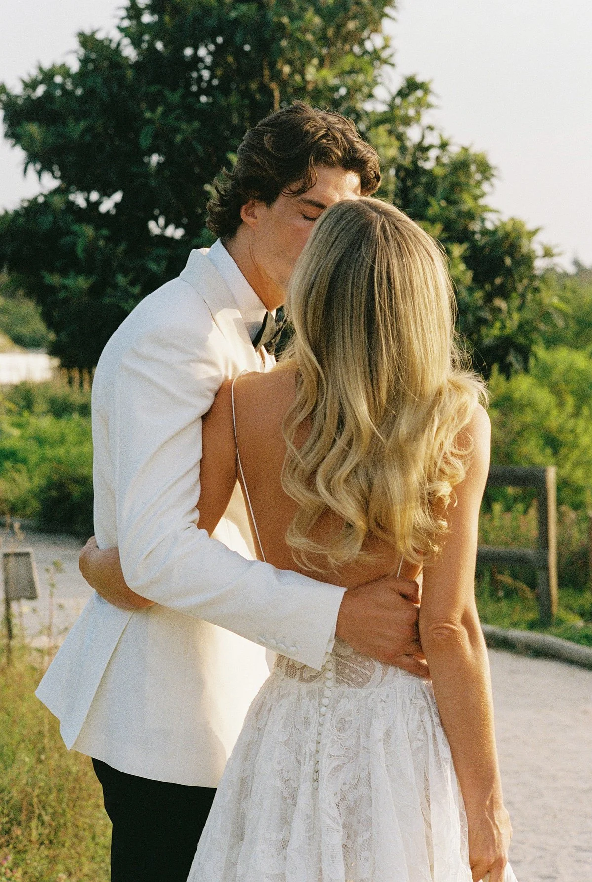 A man in a white tuxedo with a bow tie appears to be kissing a woman in a white dress with lace details, outdoors with greenery in the background.