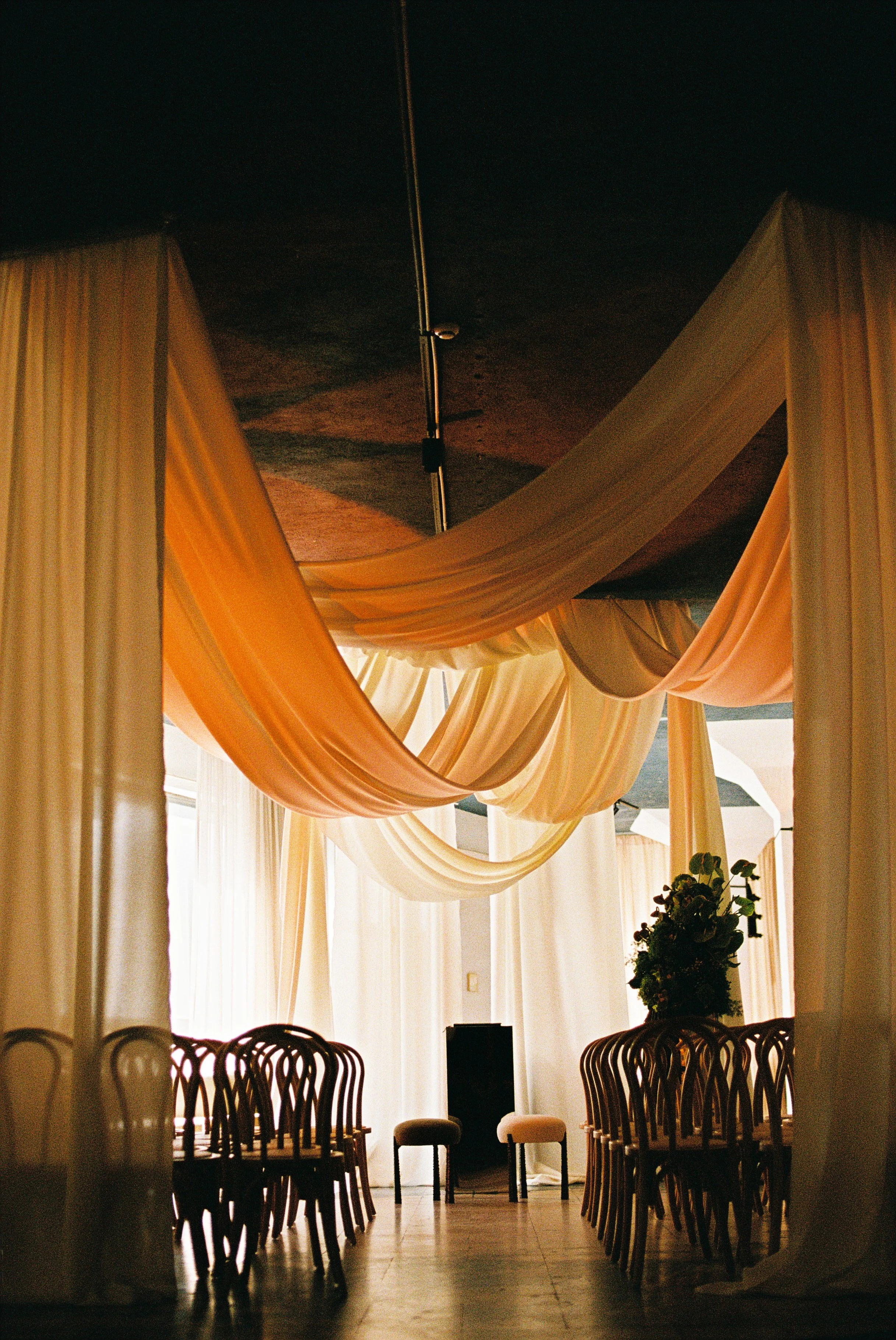 Interior of a room with ornate drapes in peach and cream hanging from a ceiling, with a chandelier above. There are dark wooden chairs arranged around the perimeter, and a large potted plant with colorful flowers near a window with sheer curtains.
