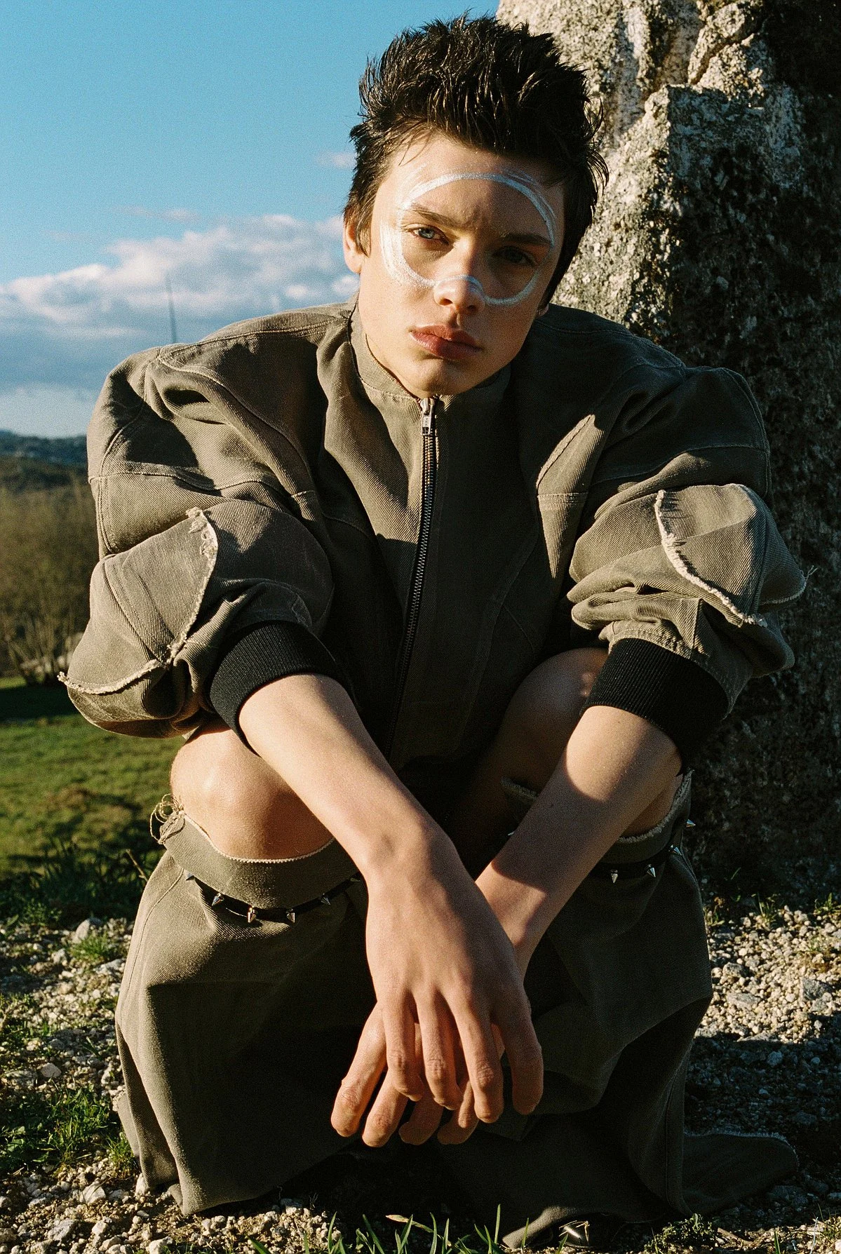 A young man with short dark hair sitting outdoors on rocky ground near a large tree with a blue sky and clouds in the background. He is wearing a khaki jacket with rolled-up sleeves, ripped khaki pants with chain details, and has white paint in a cir