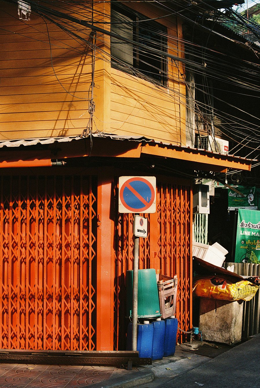 A corner building with orange-colored walls and a metal gate, with no parking signs, water containers, and a street-side stall with bags of goods.