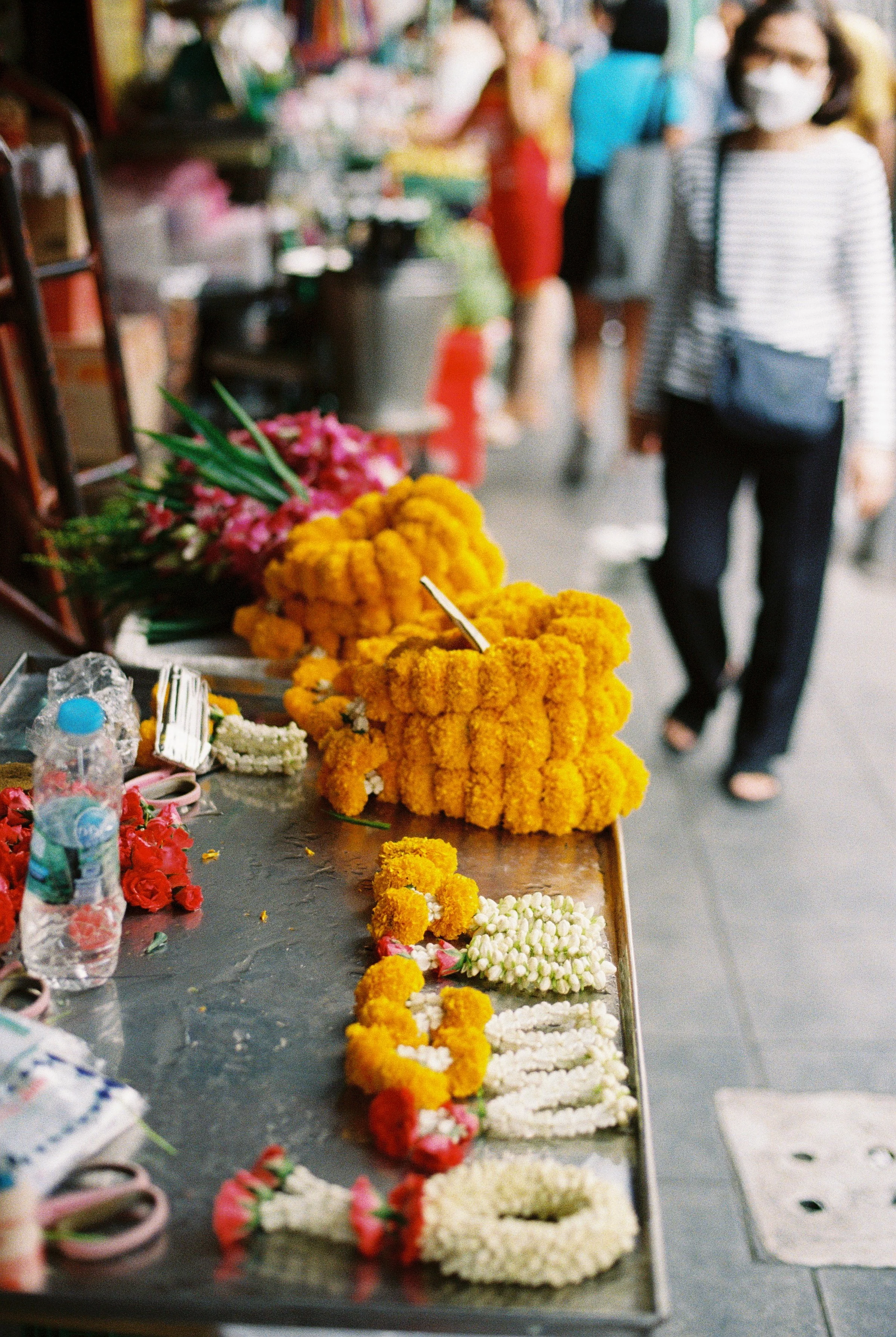 Flower arrangements made with yellow, pink, and white flowers displayed on a table at a market or event, with people in masks in the background.