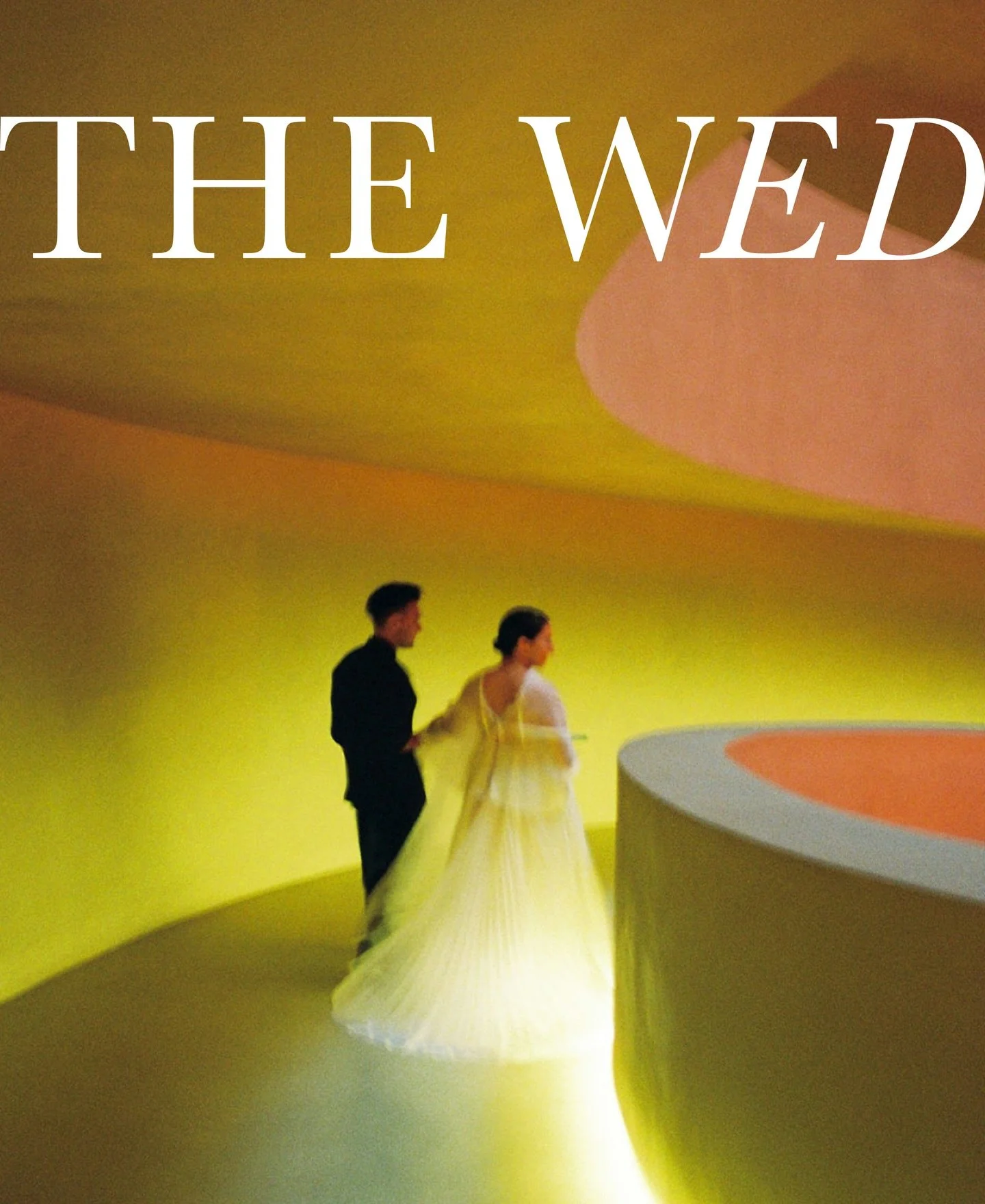 A man in a dark suit and a woman in a yellow dress stand in a yellow room with a curved ceiling and a round orange and gray table in the foreground.