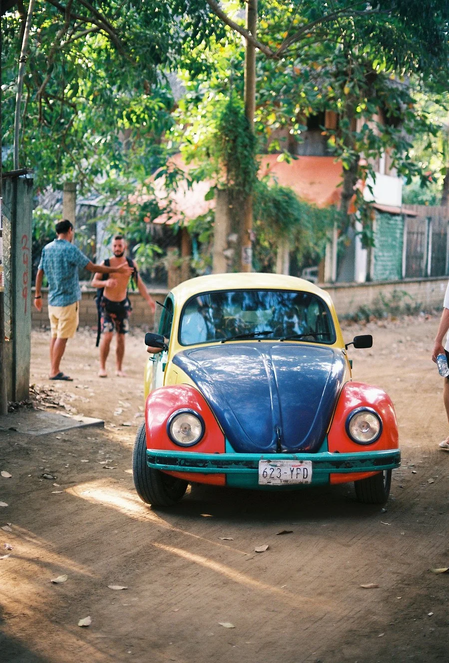A vintage Volkswagen Beetle car parked on an unpaved dirt street with a yellow roof, blue hood, and orange fenders in a leafy neighborhood. Three people are near the car, one shirtless and two dressed casually, with trees and houses in the background