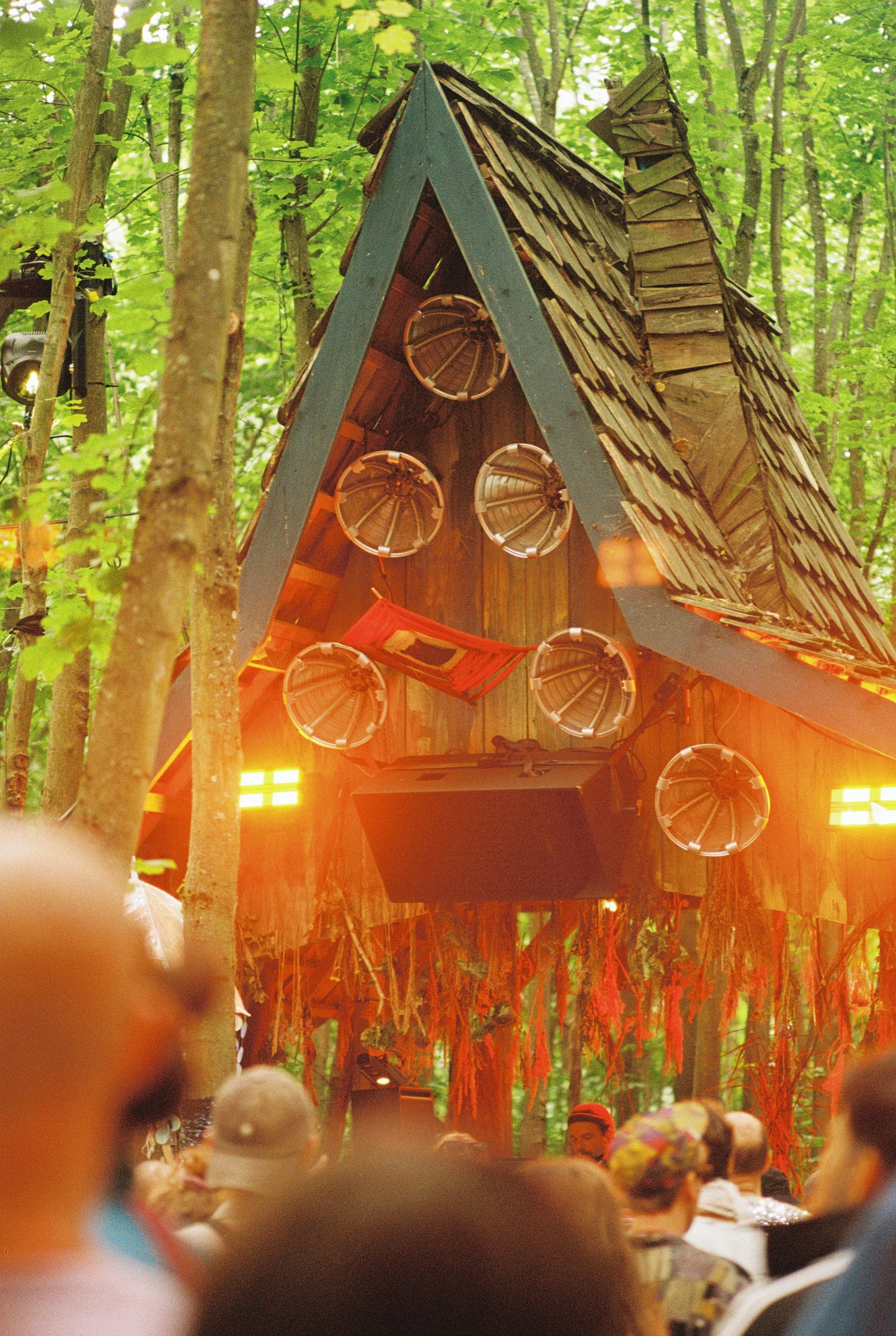 A wooden stage with a triangular roof and circular decorations hanging from it, set in a wooded area during the daytime with a large crowd of people in the foreground.