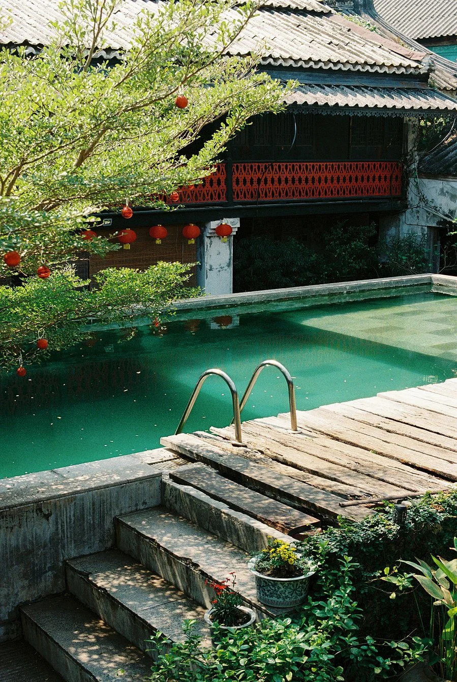 A backyard with a swimming pool, steps leading into the pool, a tree with red lanterns, and a house with a balcony and tiled roof in the background.
