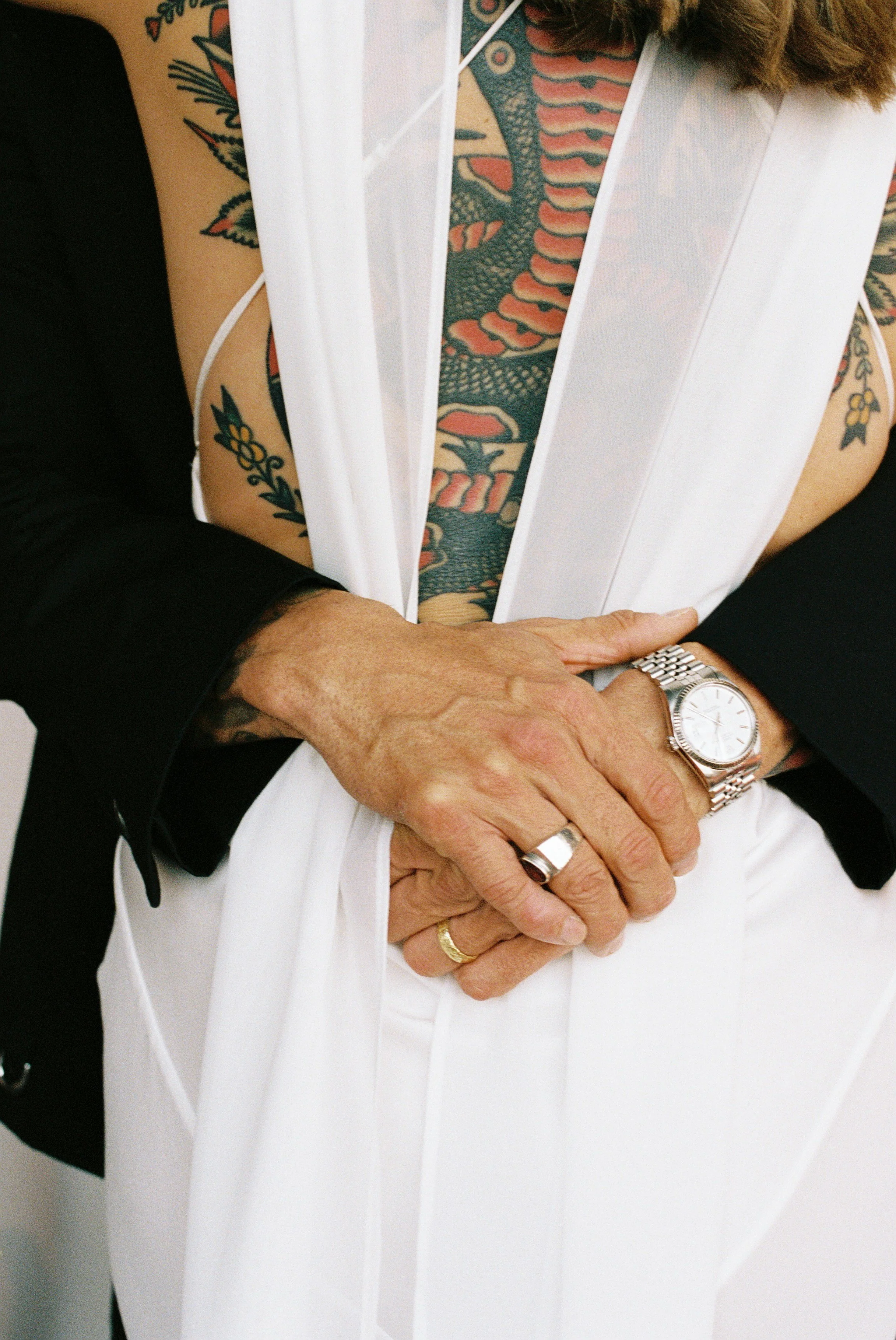 Close-up of a person with tattoos on their arm and torso, wearing a black jacket, holding a white garment or fabric with their left arm, and wearing a watch and rings on their fingers.