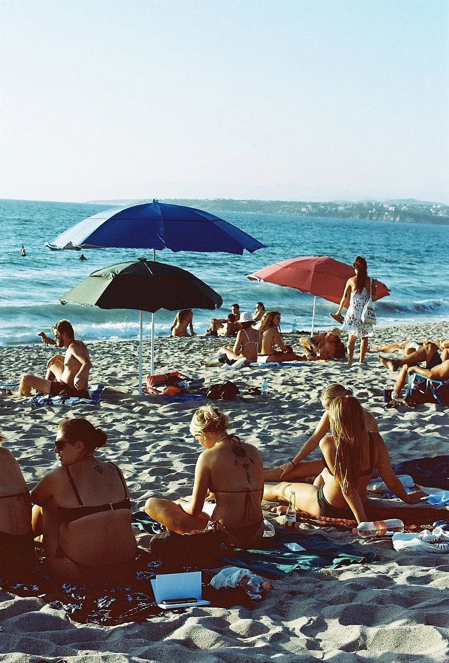 People relaxing on a sandy beach under umbrellas near the ocean, with others swimming and walking along the shoreline.