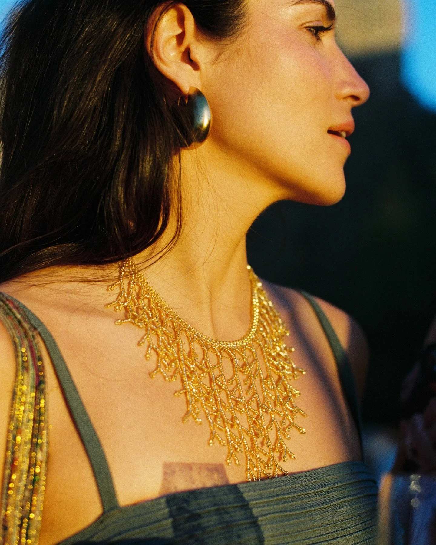 A woman with dark hair wearing gold jewelry, including a necklace and earrings, and a strapless green dress with striped detail, is captured in profile in a close-up shot.