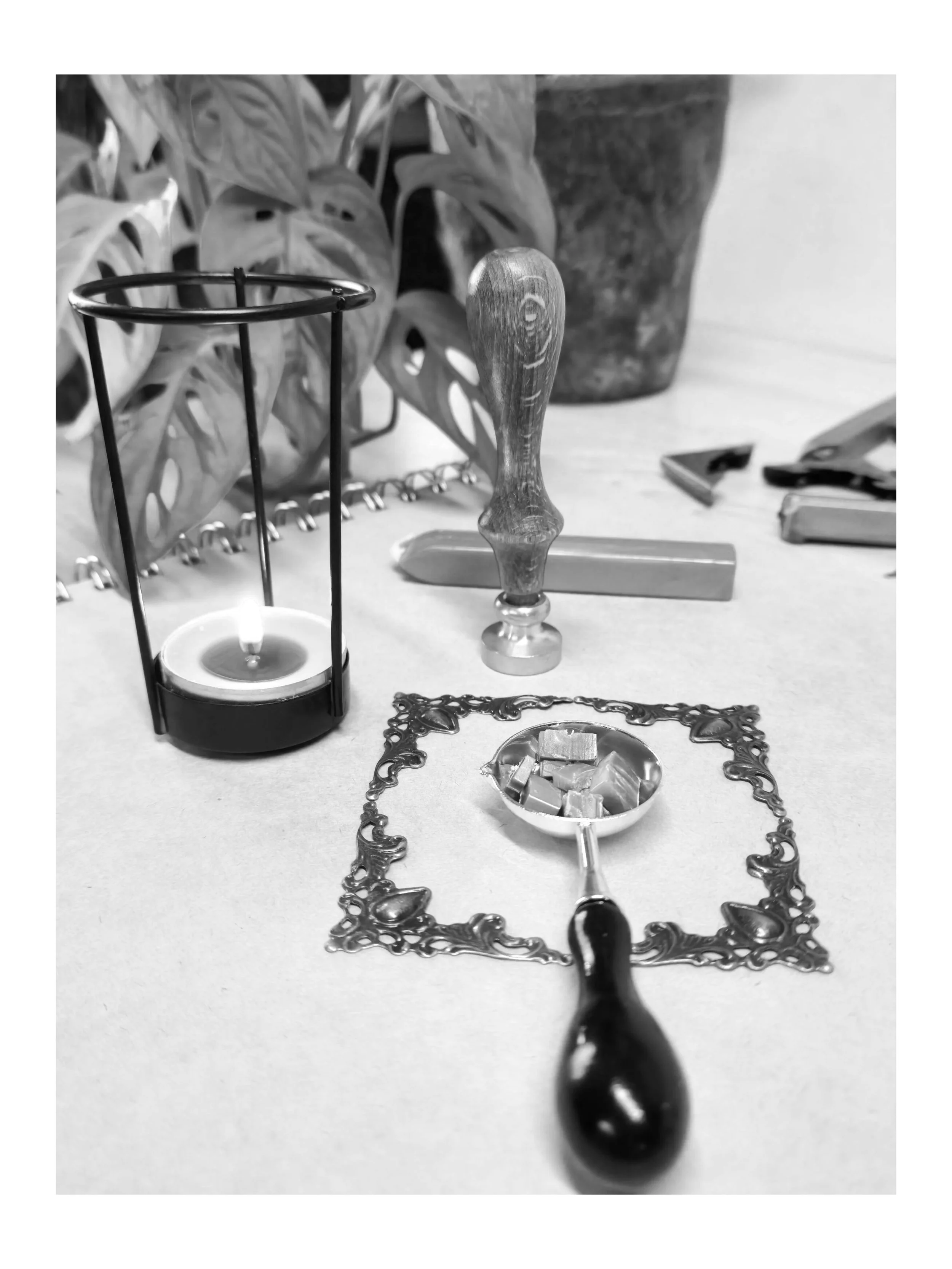 Schwarz-Weiß-Fotografie mit Utensilien für den Wachsstempel, darunter Stempel, Wachslöffel und Kerze, arrangiert auf einer Tischfläche.