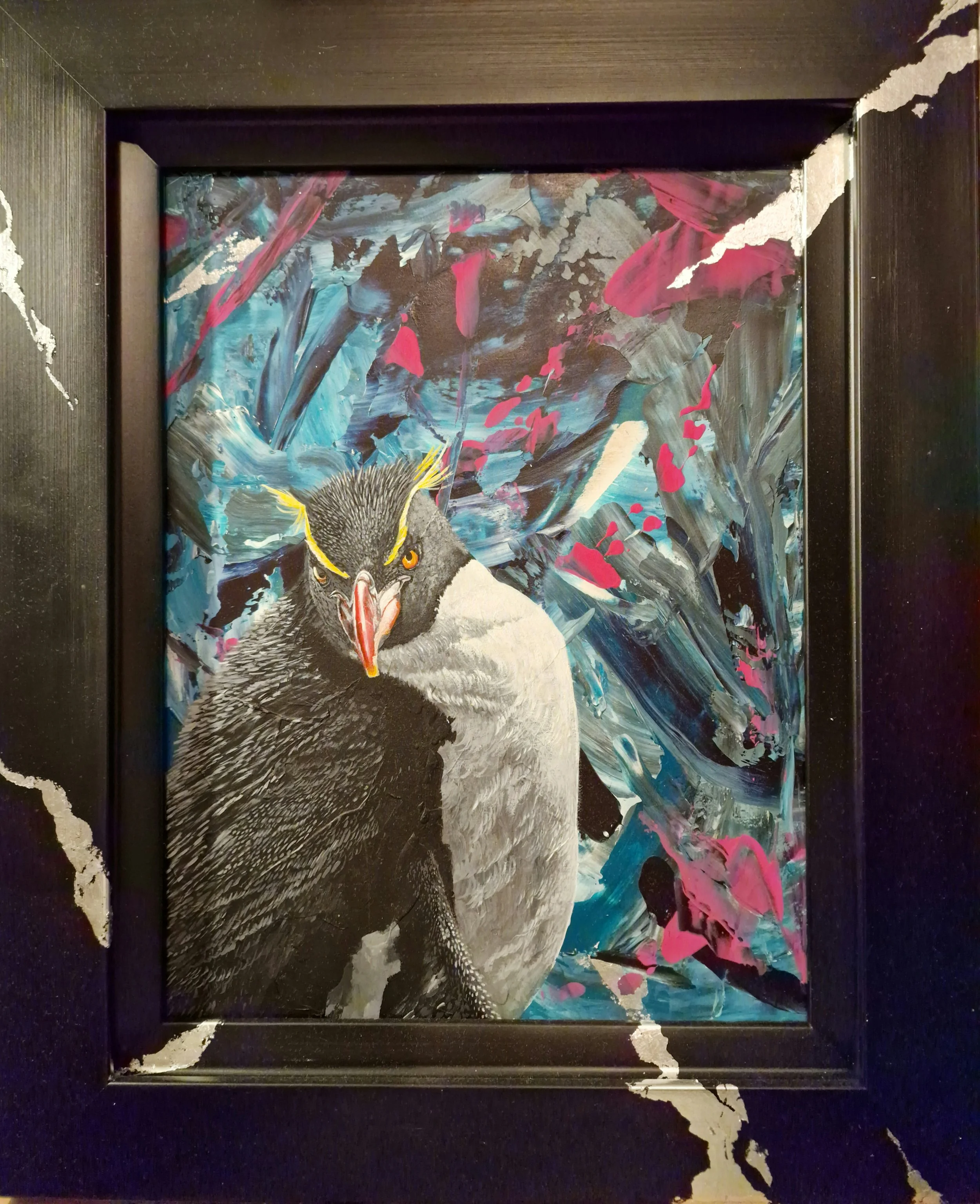 Zeitgenössisches Gemälde eines Felsenpinguin vor einem dynamischen abstrakten Hintergrund in Blau, Pink, Schwarz und Weiß, präsentiert in einem schwarzen massiven Rahmen. Schlagmetall in Silber zieht sich über Rahmen und Werk..