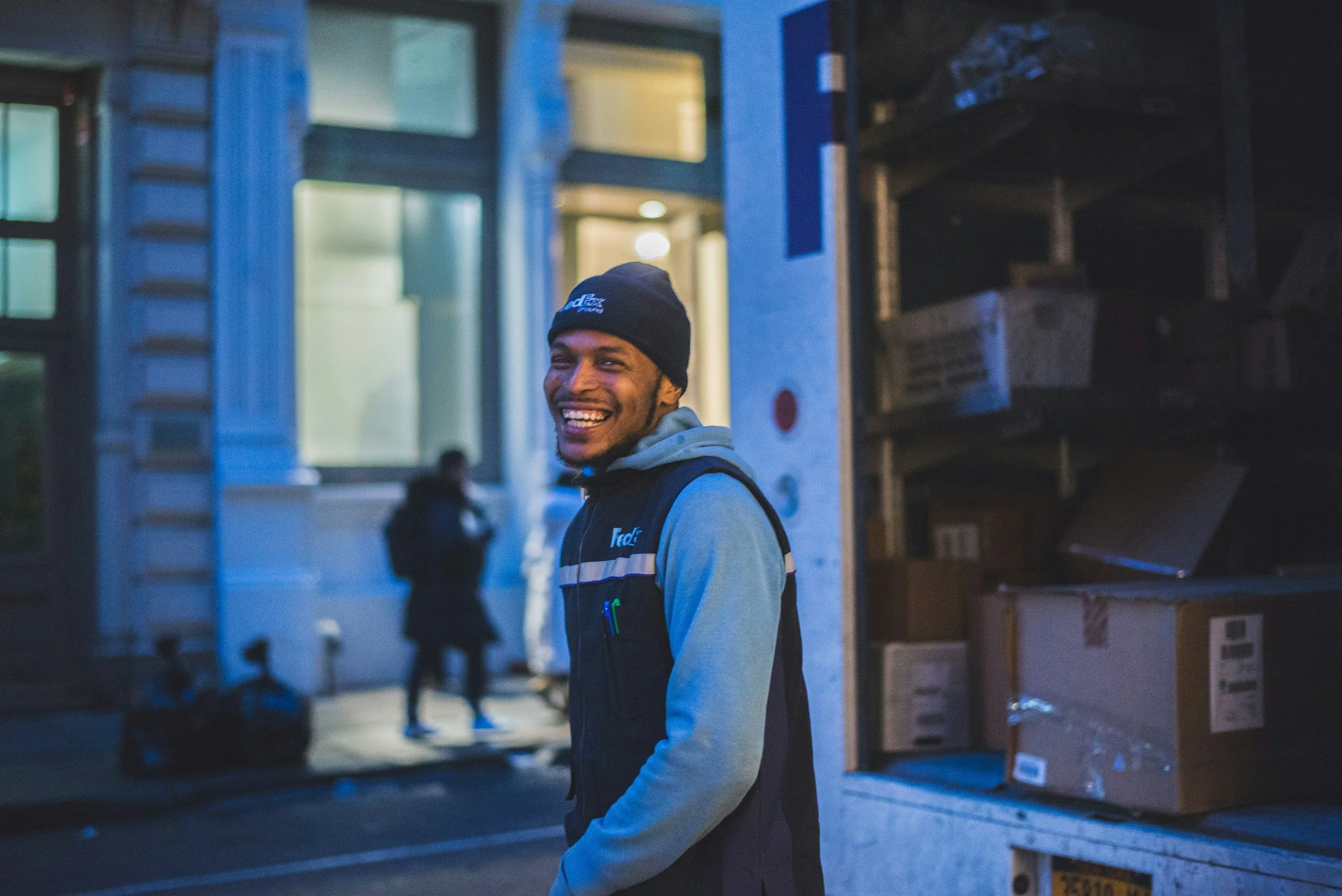 A young man smiling and wearing a dark blue beanie and gray hoodie, standing near packed shelves of cardboard boxes in a dimly lit warehouse or storage area.