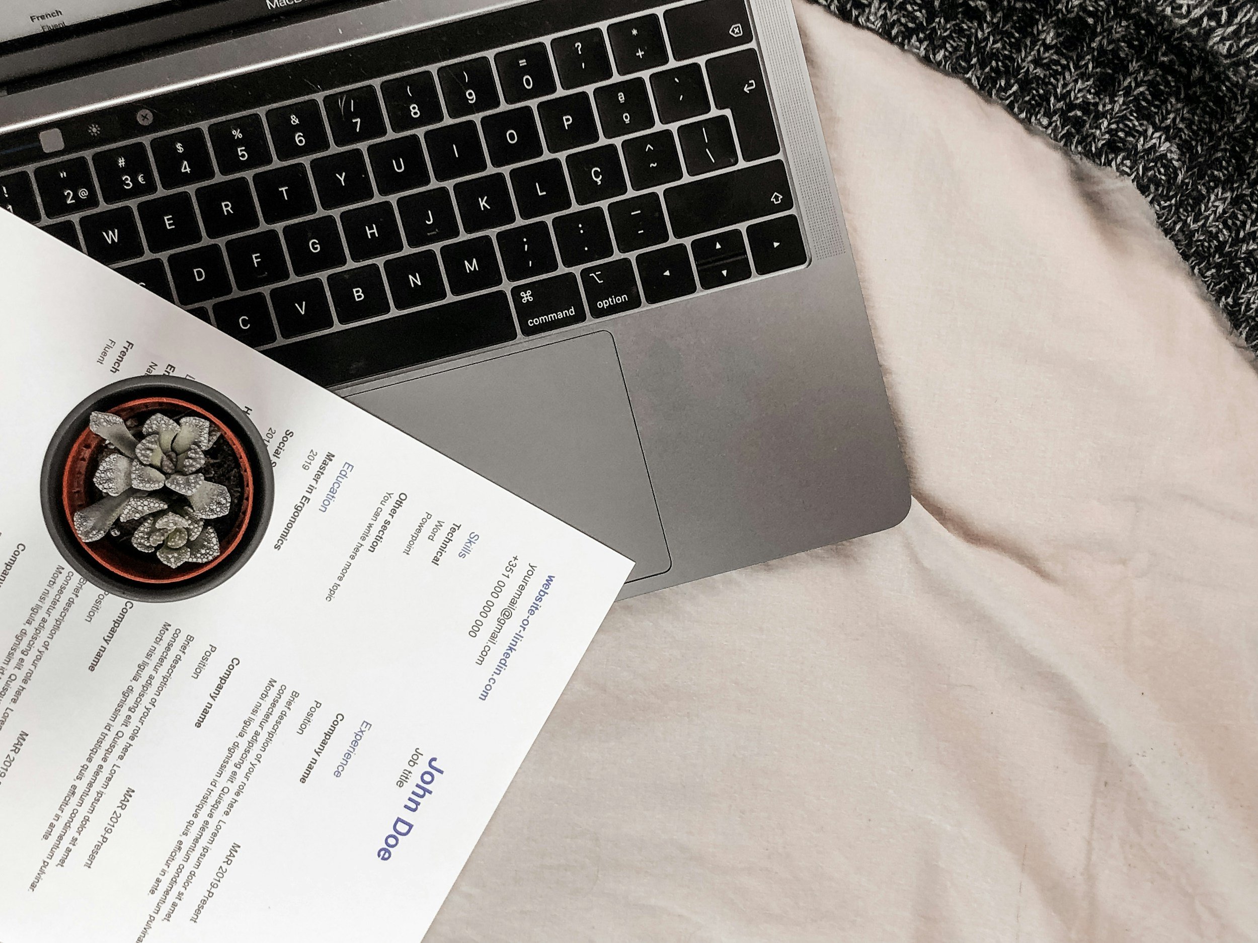 Close-up of a laptop keyboard with a portion of a printed document and a small potted succulent plant on top, resting on a soft surface with a person's sweater sleeve visible in the background.