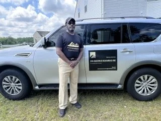 Man standing next to a silver vehicle with a black sign on the door, outdoors on grass with a building and blue sky in the background.