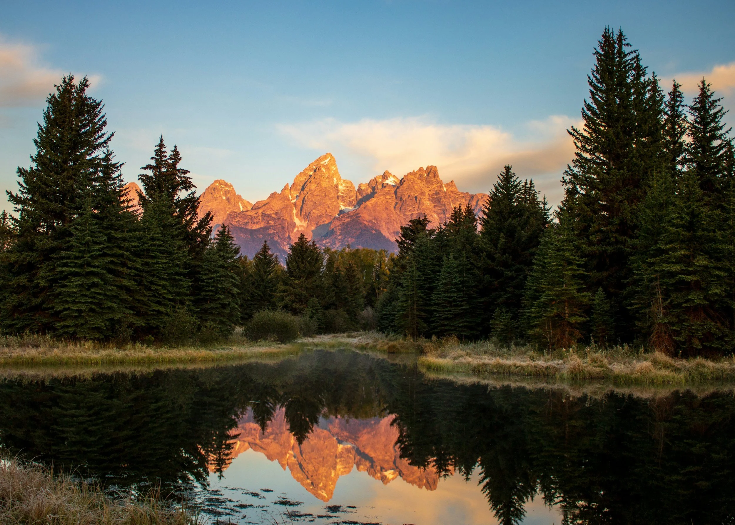 Sunset over rugged mountain peaks reflected in a calm lake, surrounded by evergreen trees.