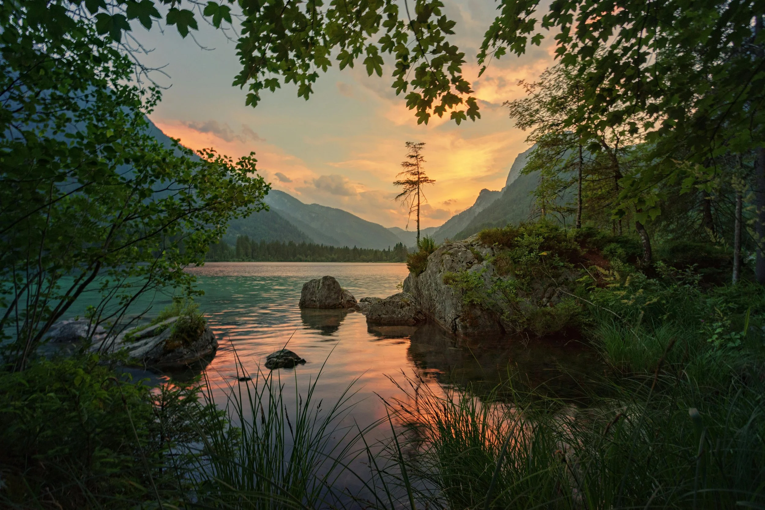 A scenic view of a river surrounded by trees and mountains during sunset, with the sky painted in orange and pink hues.