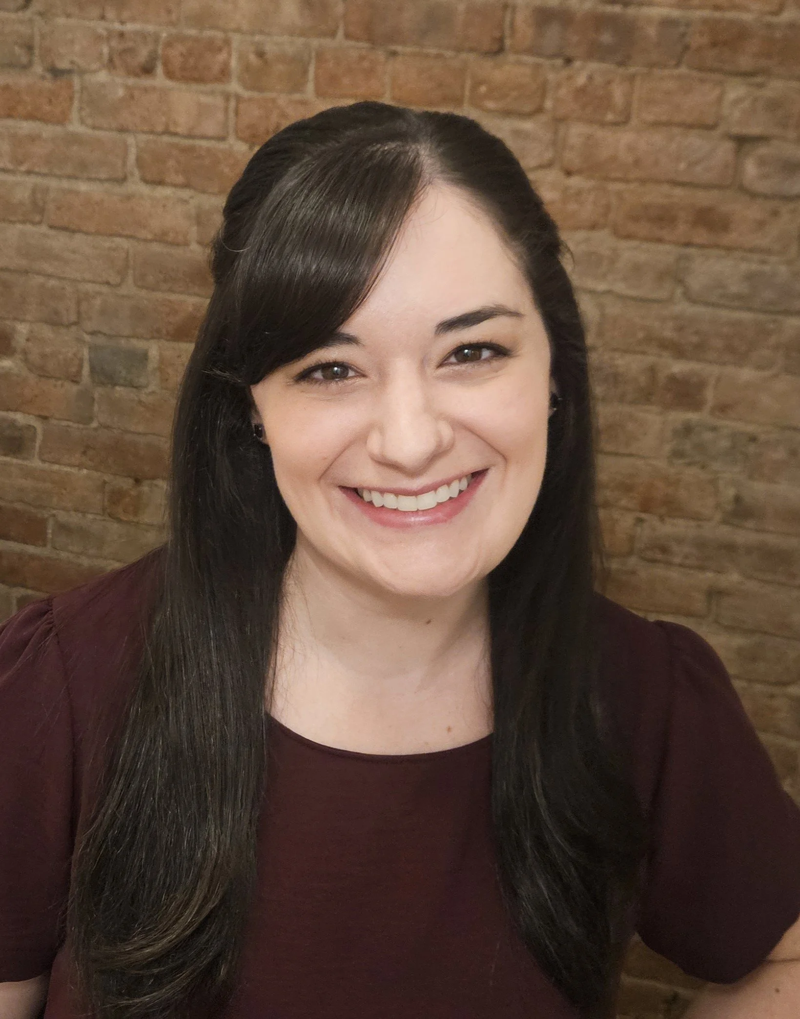A woman with long dark hair smiling against a brick wall background.