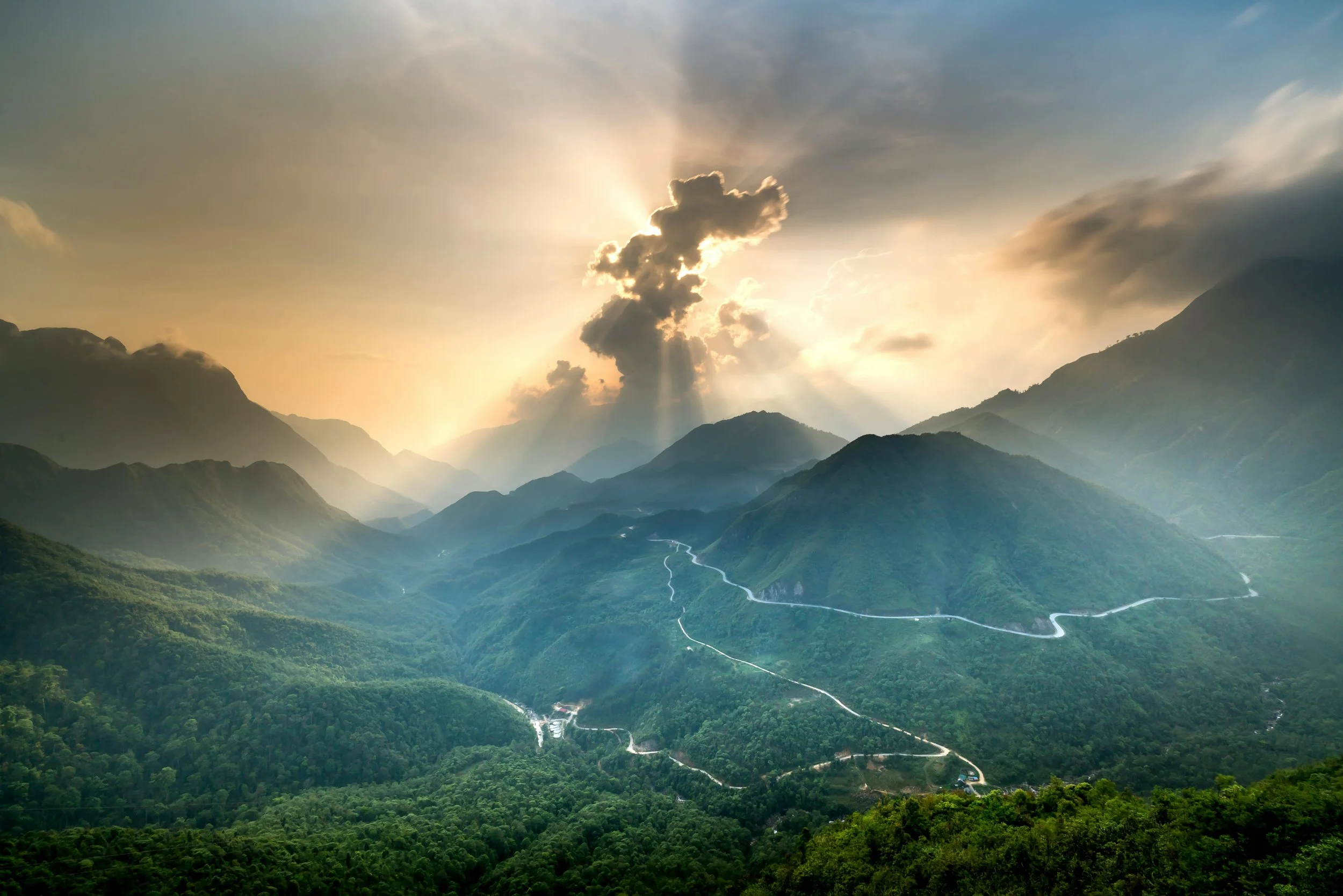 Scenic view of a mountain landscape during sunrise or sunset with a winding road through lush green hills and a cloud formation with sun rays shining through.