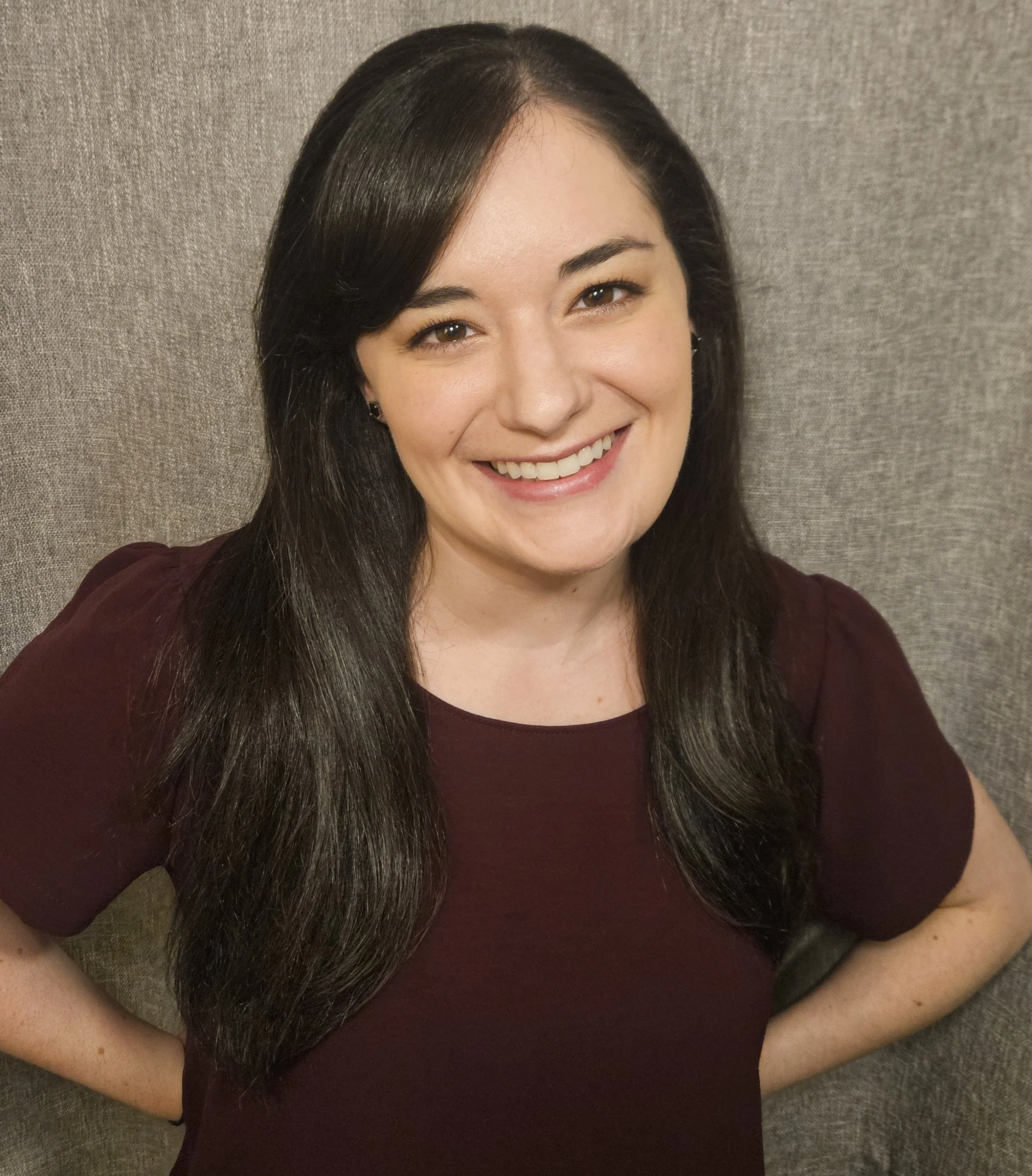 A woman with long dark hair, smiling, sitting on a gray textured surface, wearing a dark maroon top.