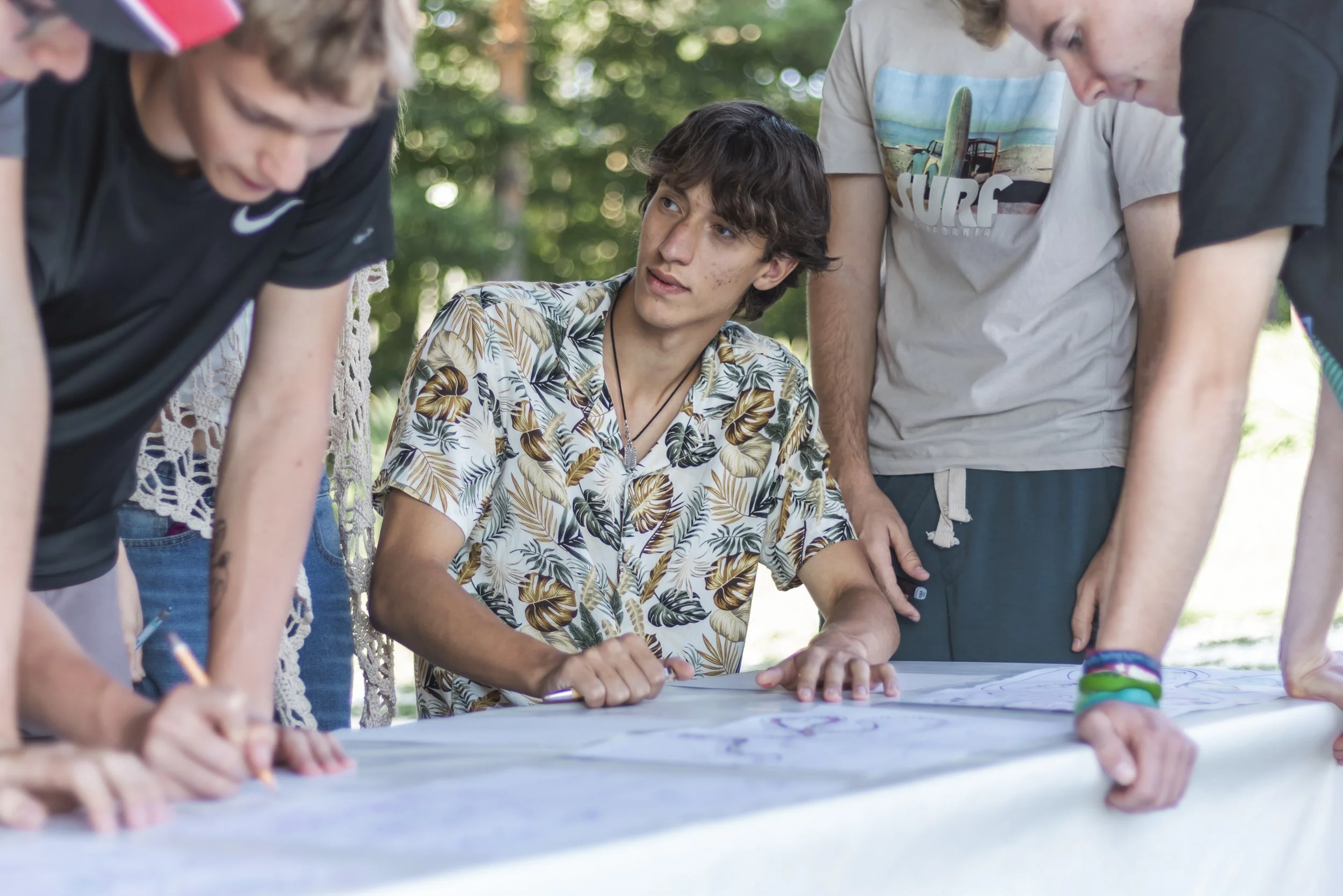 A group of teenagers gathered around a table outdoors working on a large piece of paper or map, with trees and greenery in the background.