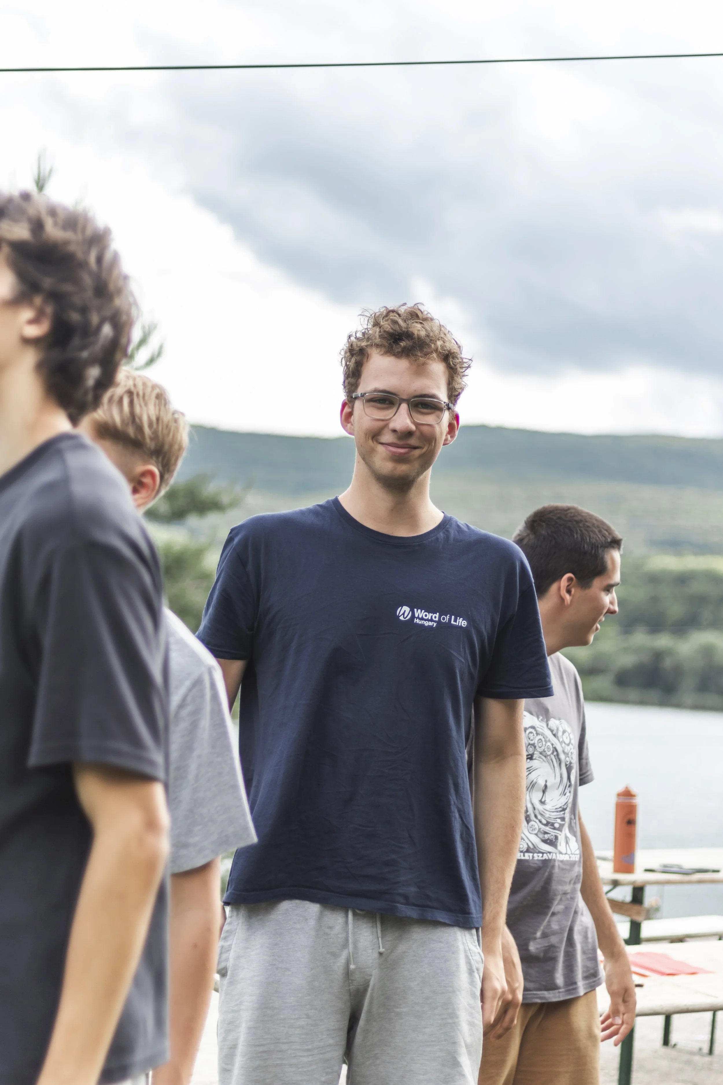 A group of young men outdoors, standing next to each other, with a scenic mountain lake background. The man in the center is smiling and wearing glasses, a navy blue T-shirt with 'Word of Life Hungary' printed on it, and gray sweatpants.