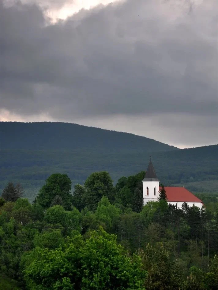 A small white church with a pointed steeple surrounded by green trees, with dark mountain hills and a cloudy sky in the background.