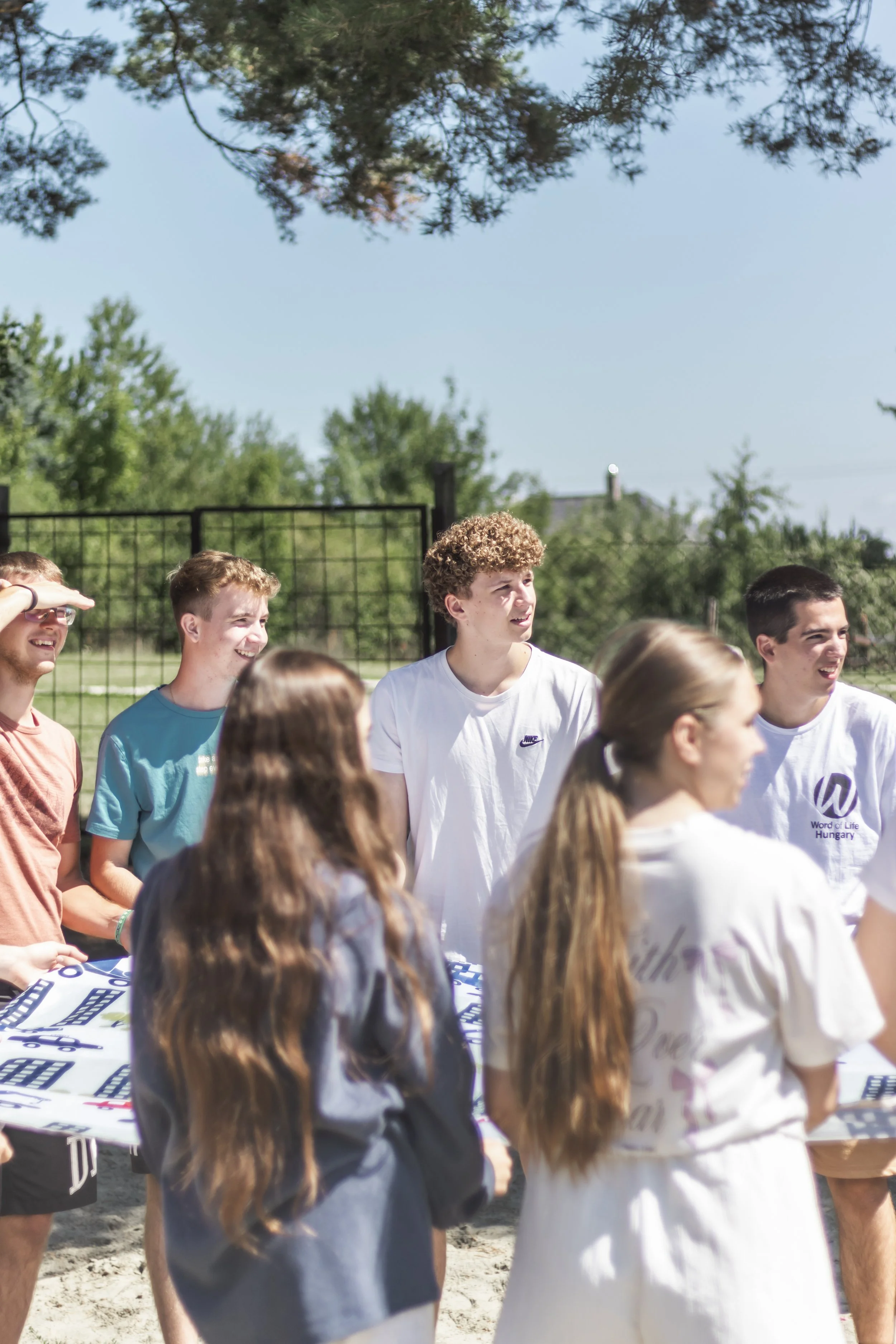 Group of young people gathered outdoors on a sunny day, some wearing casual t-shirts. The background features a black fence and trees. The scene appears to be a communal or event setting.