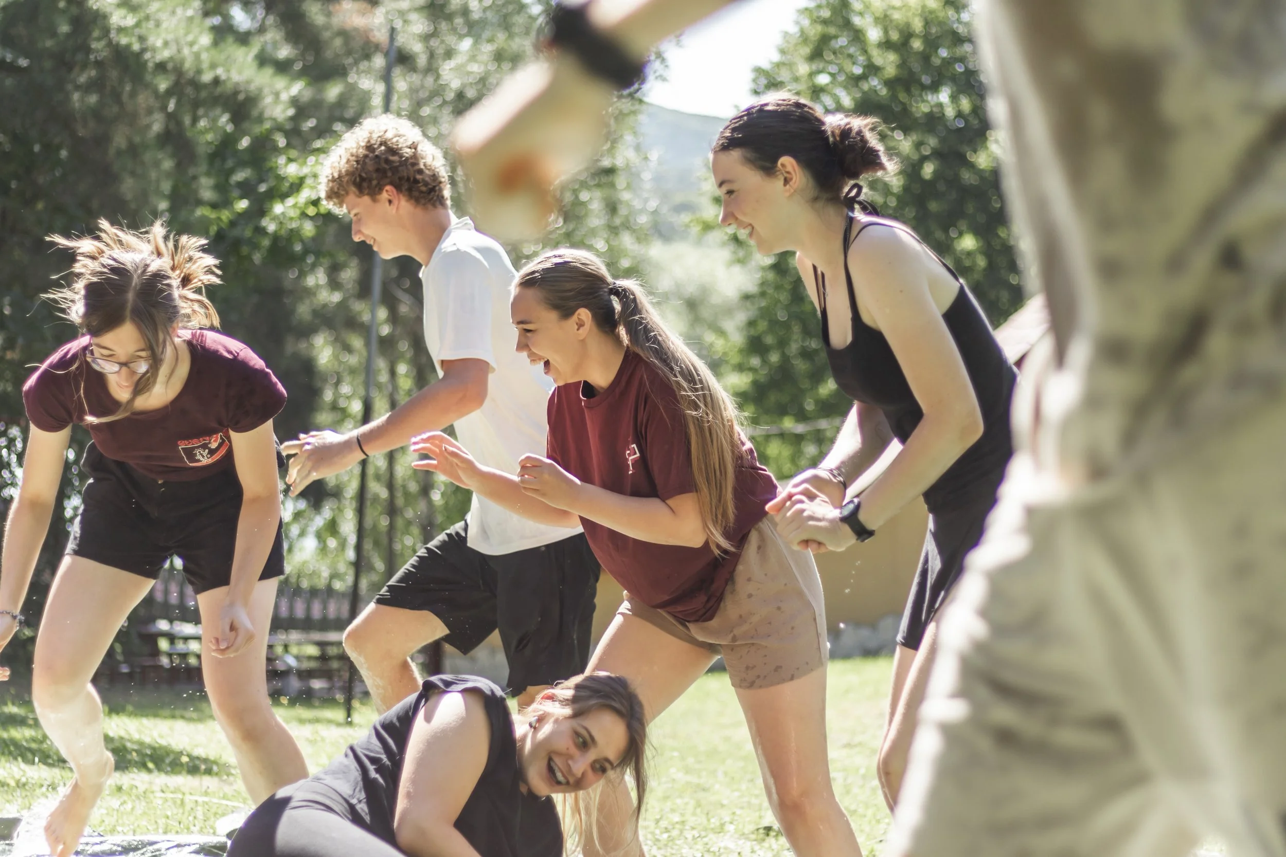 Group of young people participating in an outdoor team-building activity, stacked in a human pyramid, smiling and laughing in a park with sunlight filtering through trees.