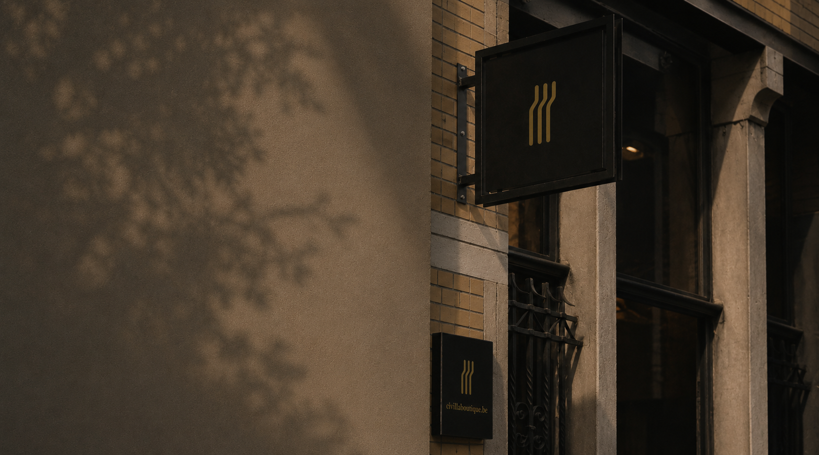 Exterior of a building with two black signs featuring a gold logo and website, one mounted on the wall and the other hanging, with brick and stucco walls, large windows, and wrought iron bars, during evening lighting.