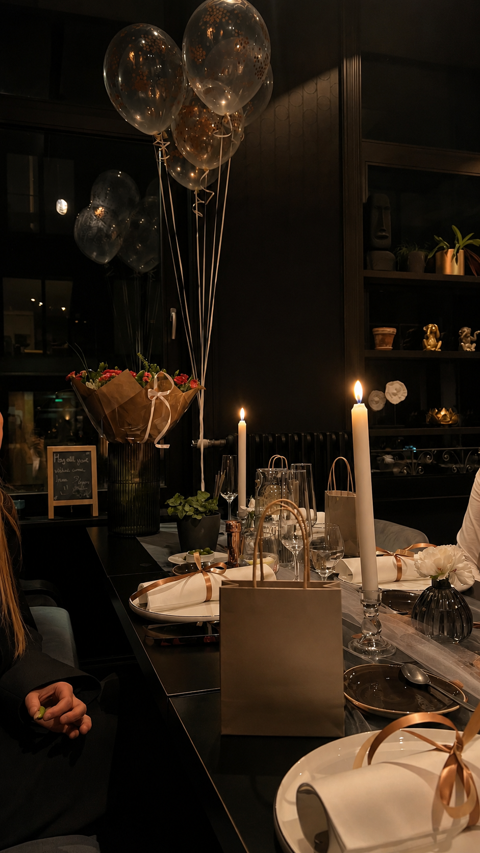 A dinner table decorated with candles, flowers, gift bags, and balloons in a dimly lit room, likely set for a celebration or special occasion.