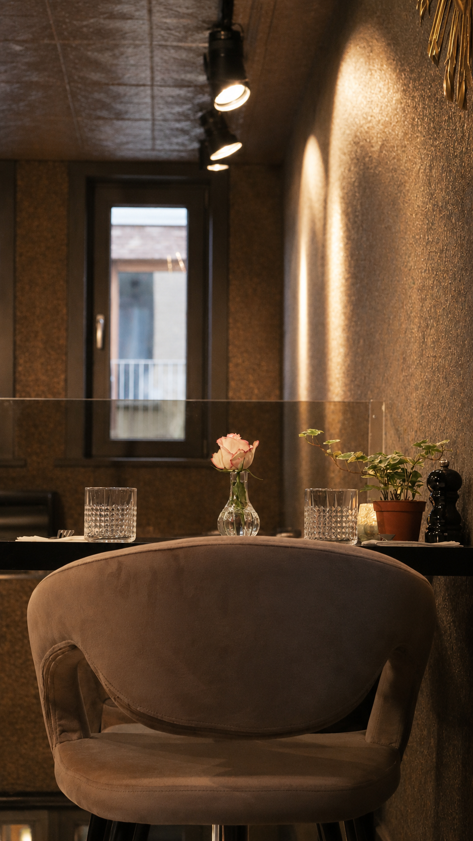 A cozy dining nook with a velvet-backed chair, a small table set with water glasses, a pink rose in a glass vase, a potted plant, and a candle, illuminated by directed ceiling lights near a window.