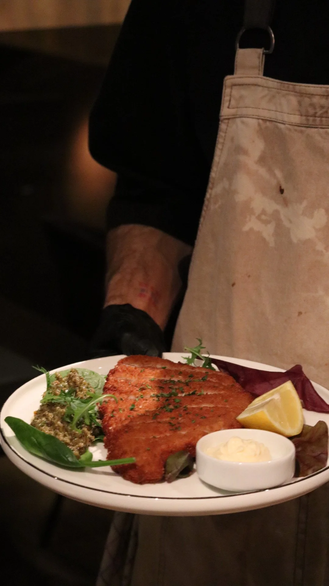 A plate of cooked salmon, garnished with herbs, served with lemon, a small bowl of tartar sauce, and a side of leafy greens and dressing, on a table with a person wearing a black shirt and a beige apron in the background.