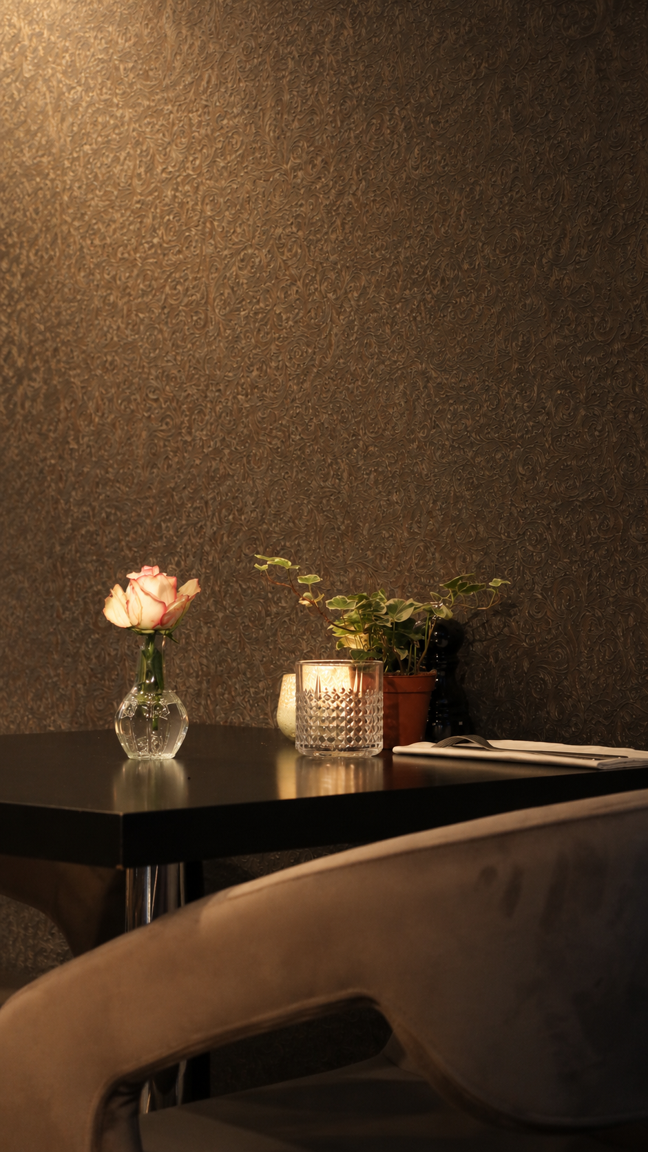 A table with a pink rose in a glass vase, a lit candle in a textured glass holder, an open magazine, and a potted plant against a textured dark wall.