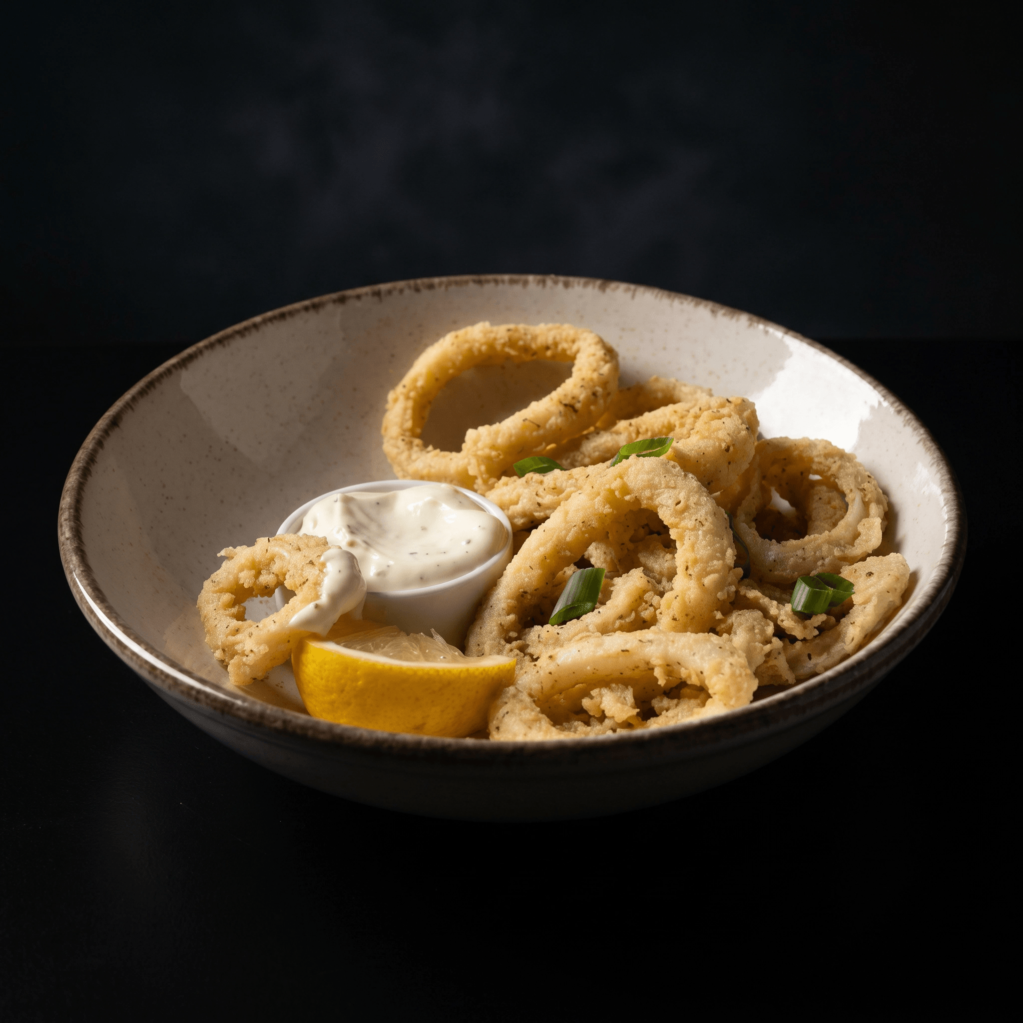 Close-up of a bowl of fried calamari rings garnished with chopped green onions and served with a dipping sauce, on a dark background.