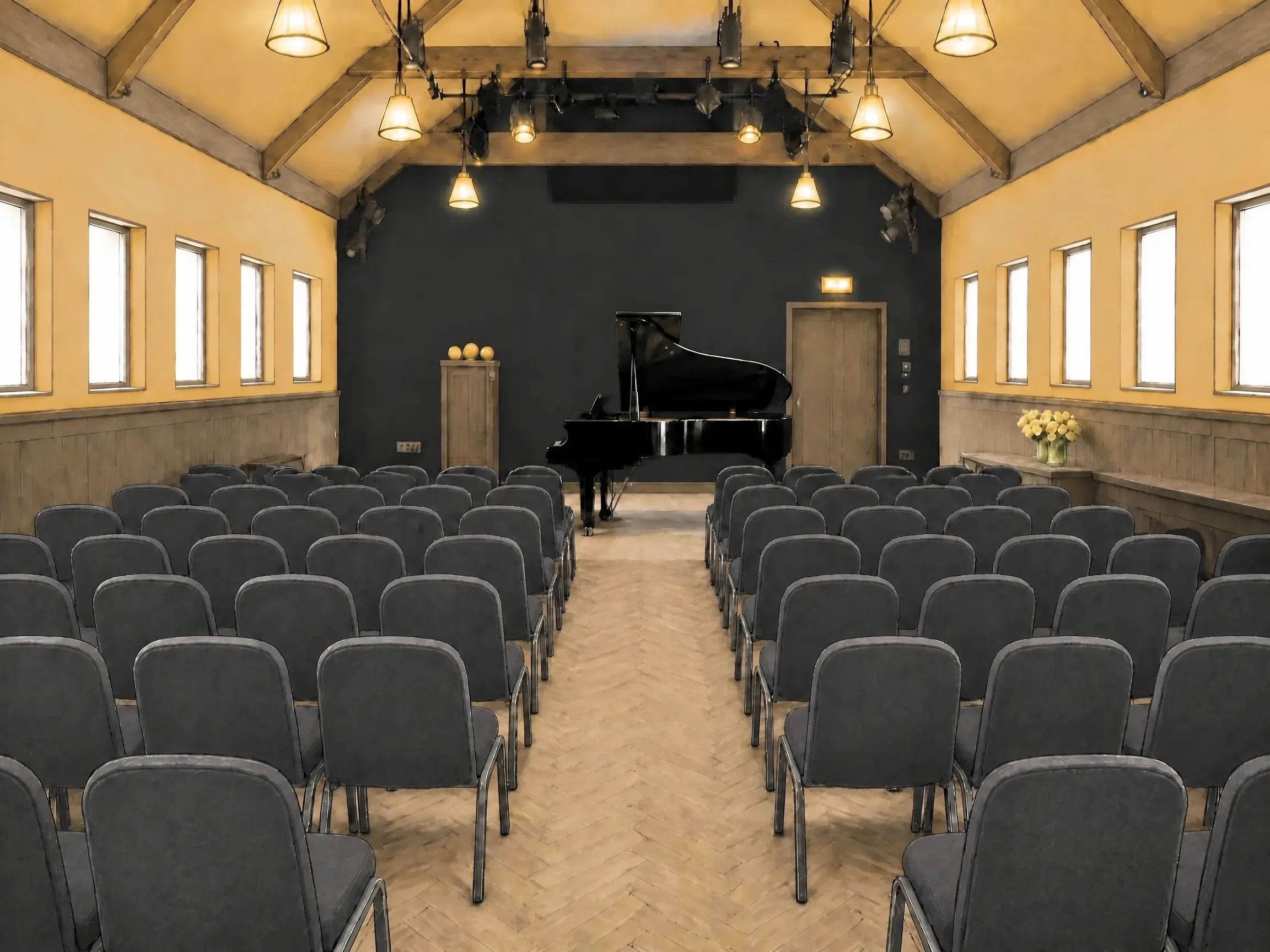 Empty room with rows of black chairs, a black grand piano, yellow walls with multiple small windows, and a stage with lighting equipment.