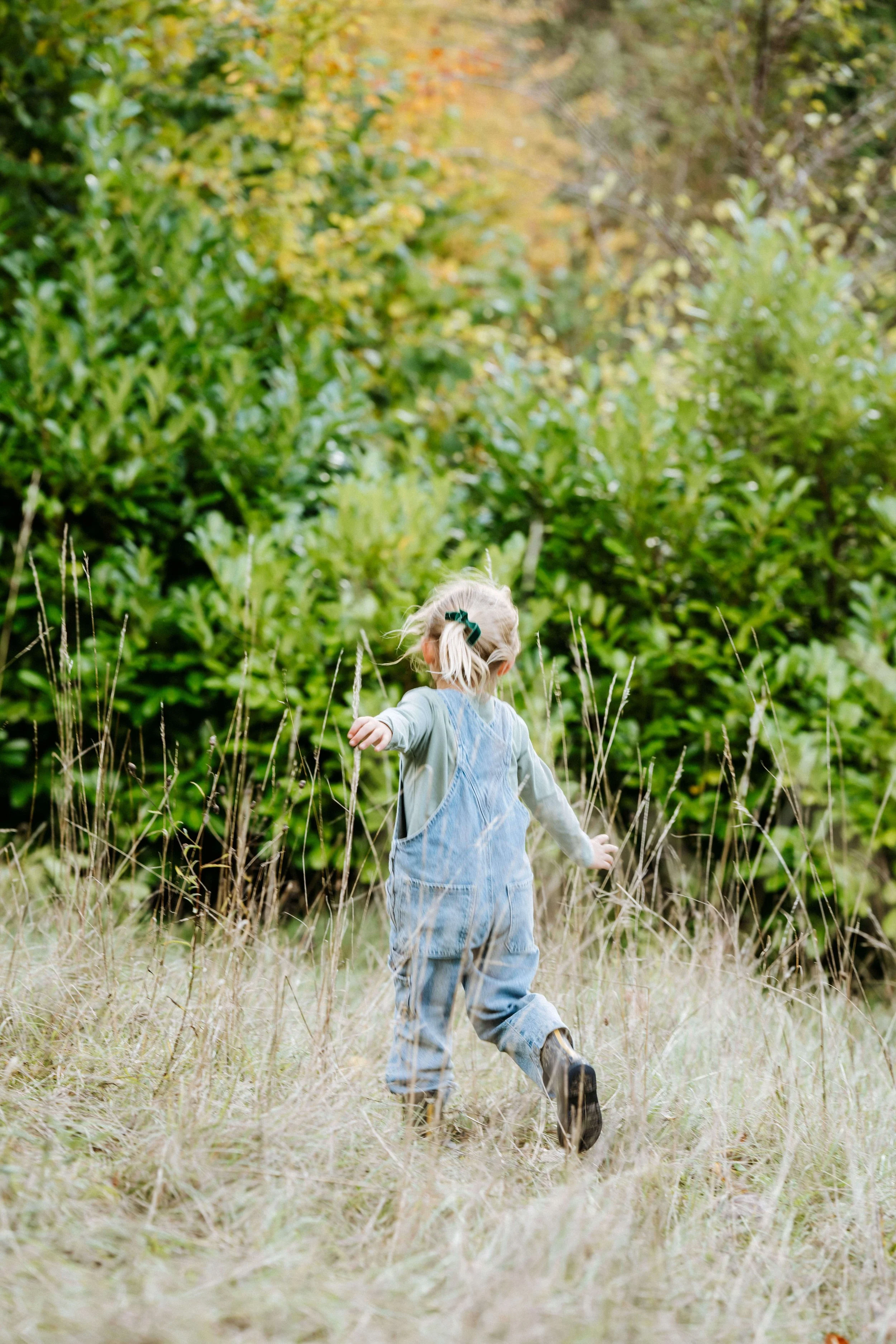 Young child running through a field of tall grass with trees and shrubbery in the background, wearing overalls and boots.