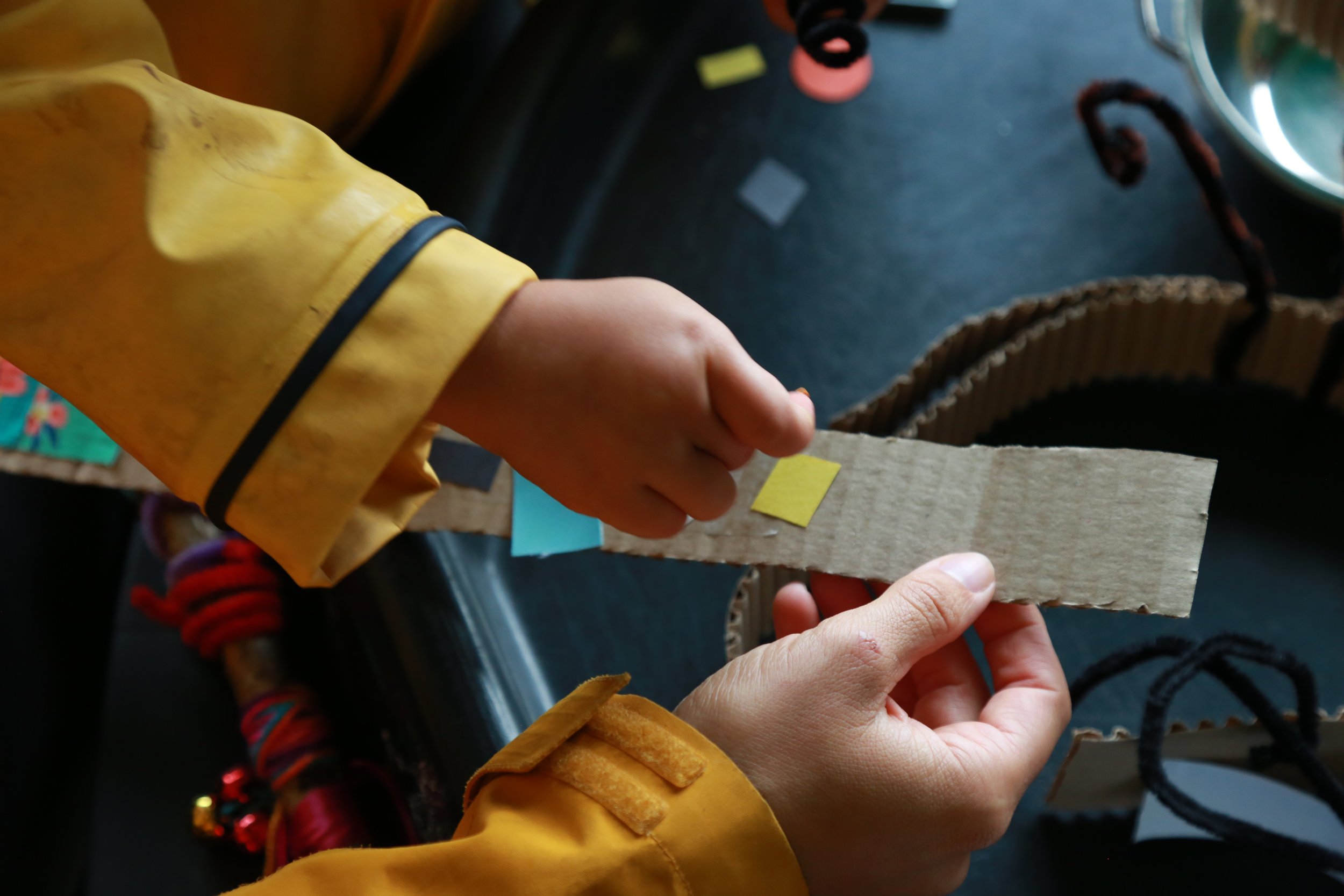 Person in a yellow jacket holding a piece of corrugated cardboard with colored tape. Other hands and objects are visible on the table.