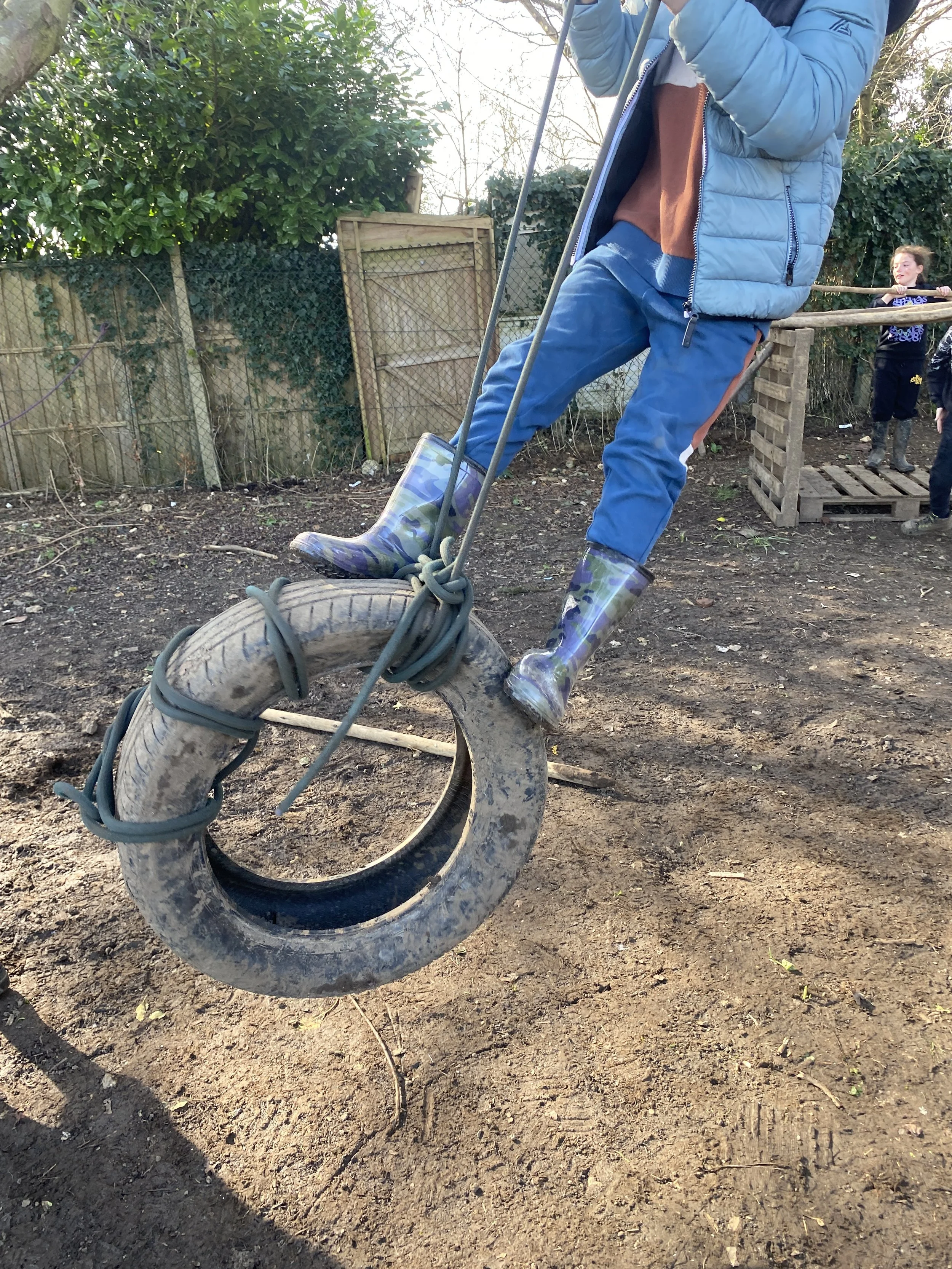 Child wearing a blue jacket and rainbow-patterned rain boots swinging on a tire swing by holding a rope, with another child holding a wooden bar in the background.