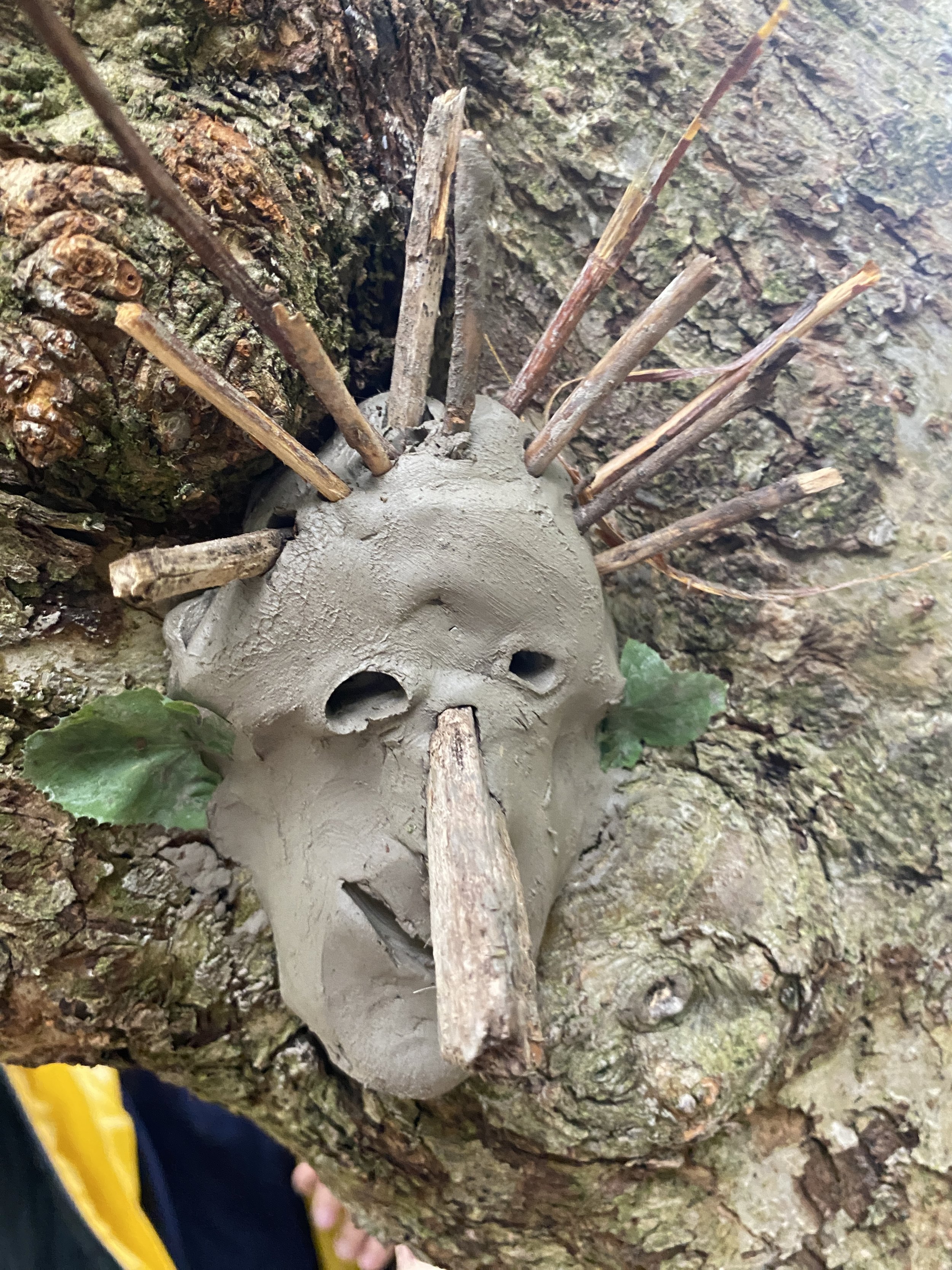 A clay animal sculpture resembling a pig, with holes for eyes and a snout, is placed on a tree trunk surrounded by bark and moss. Several small sticks are inserted into the sculpture's head.