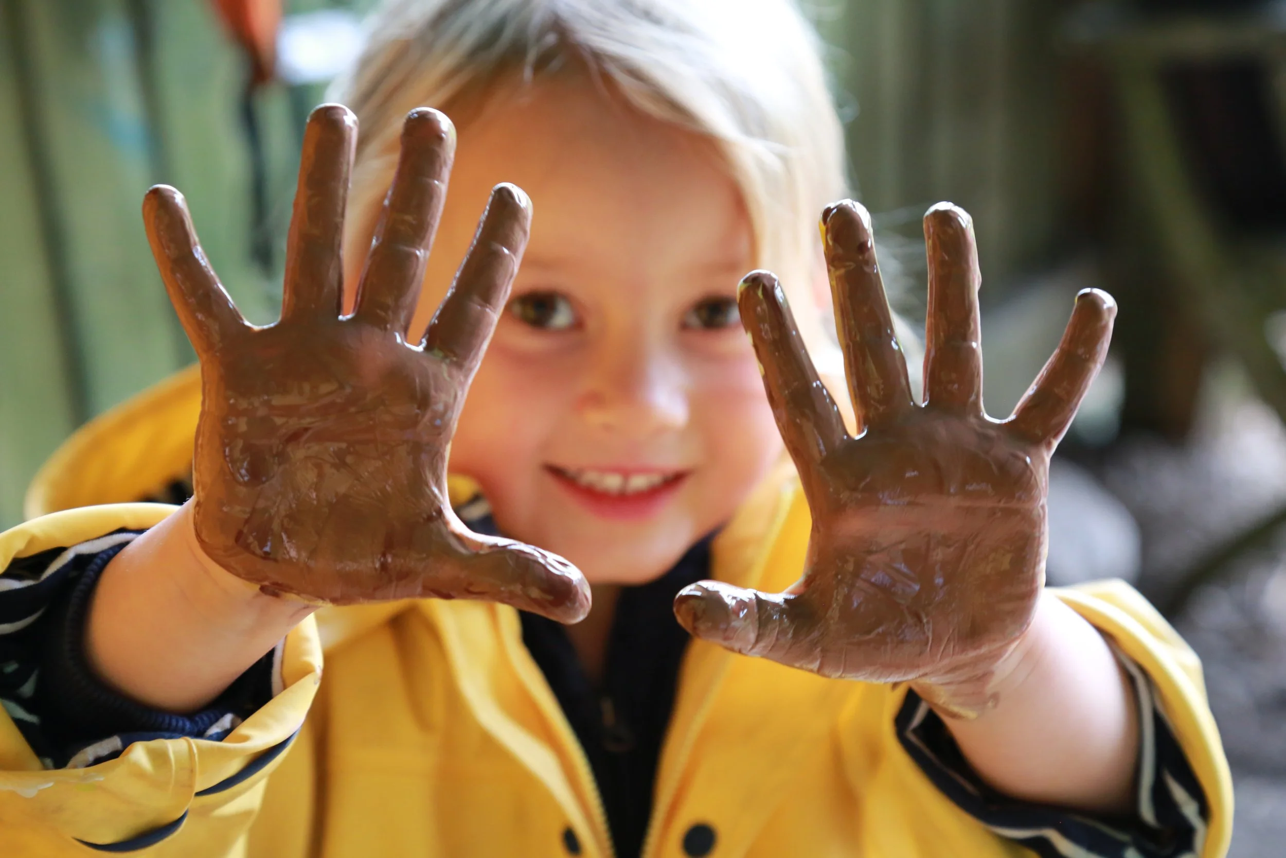 A young girl with blonde hair smiling and holding her hands up covered in chocolate while looking at the camera.