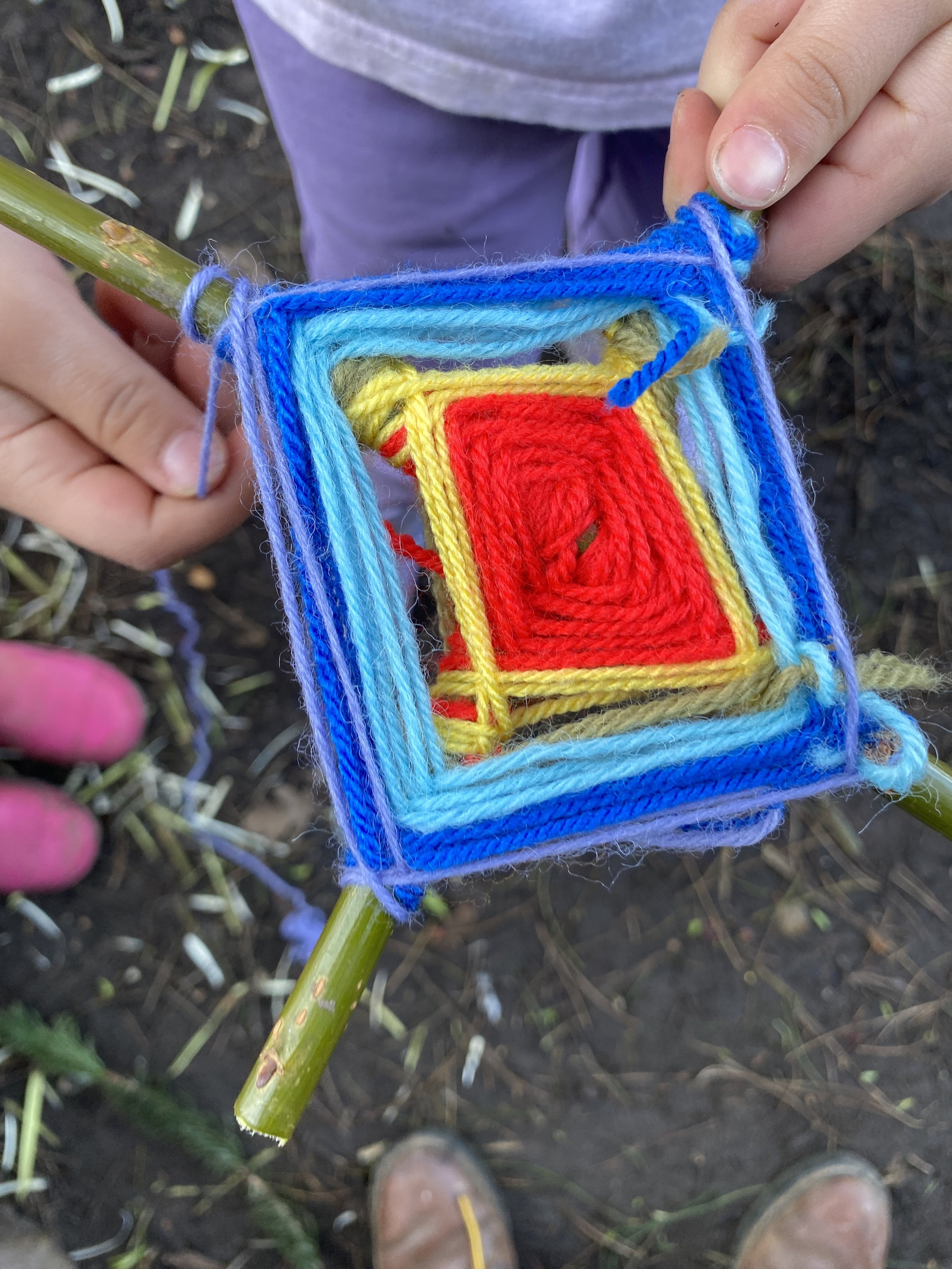 Close-up of a person weaving a colorful rainbow yarn square onto a bamboo stick, with the person's pinkish toes visible at the bottom of the image and damp soil in the background.