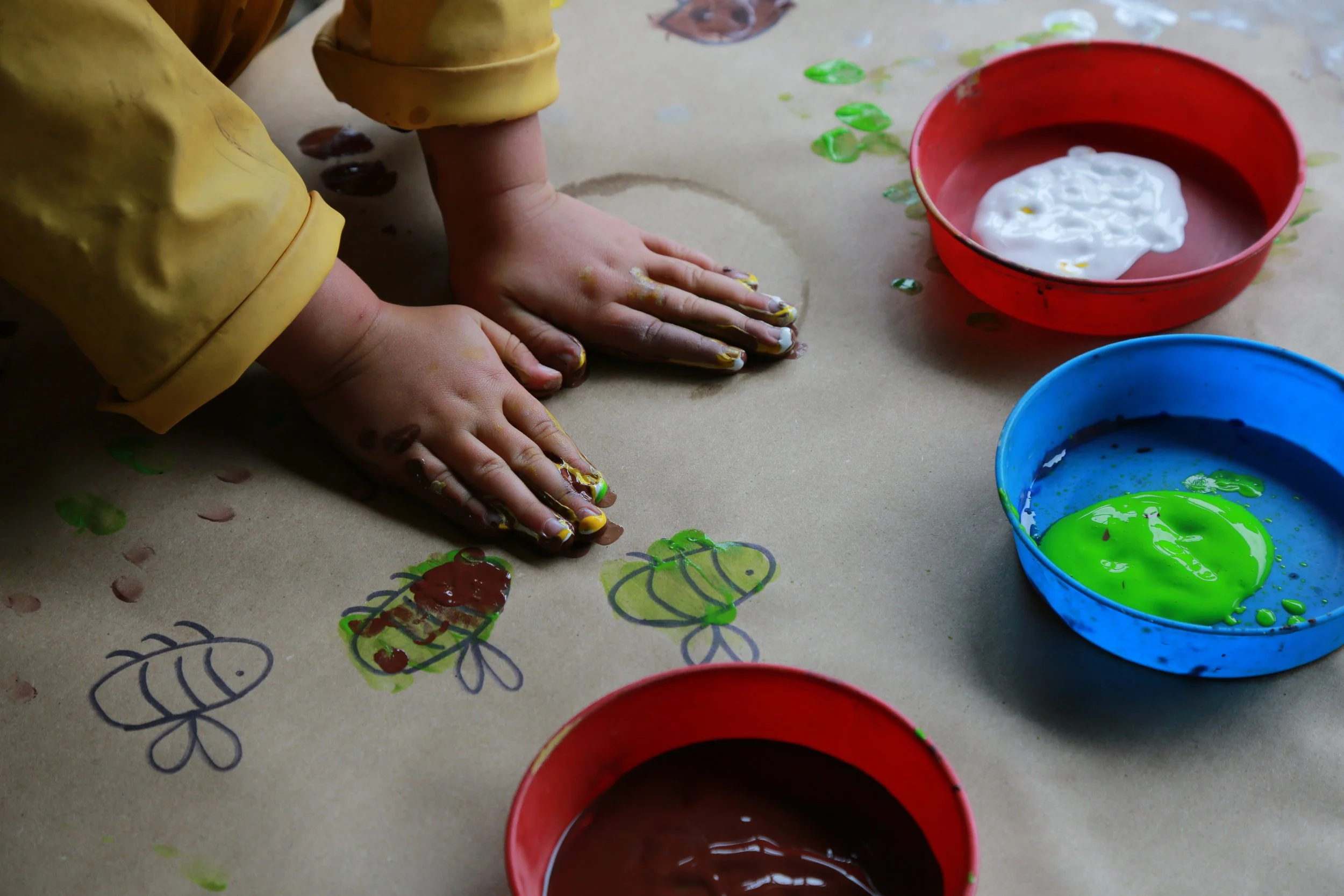 Child's hands and forearms are covered with mix of yellow and green paint, pressed flat onto a paper with colorful drawings and doodles, with bowls of red, blue, and white paint nearby.