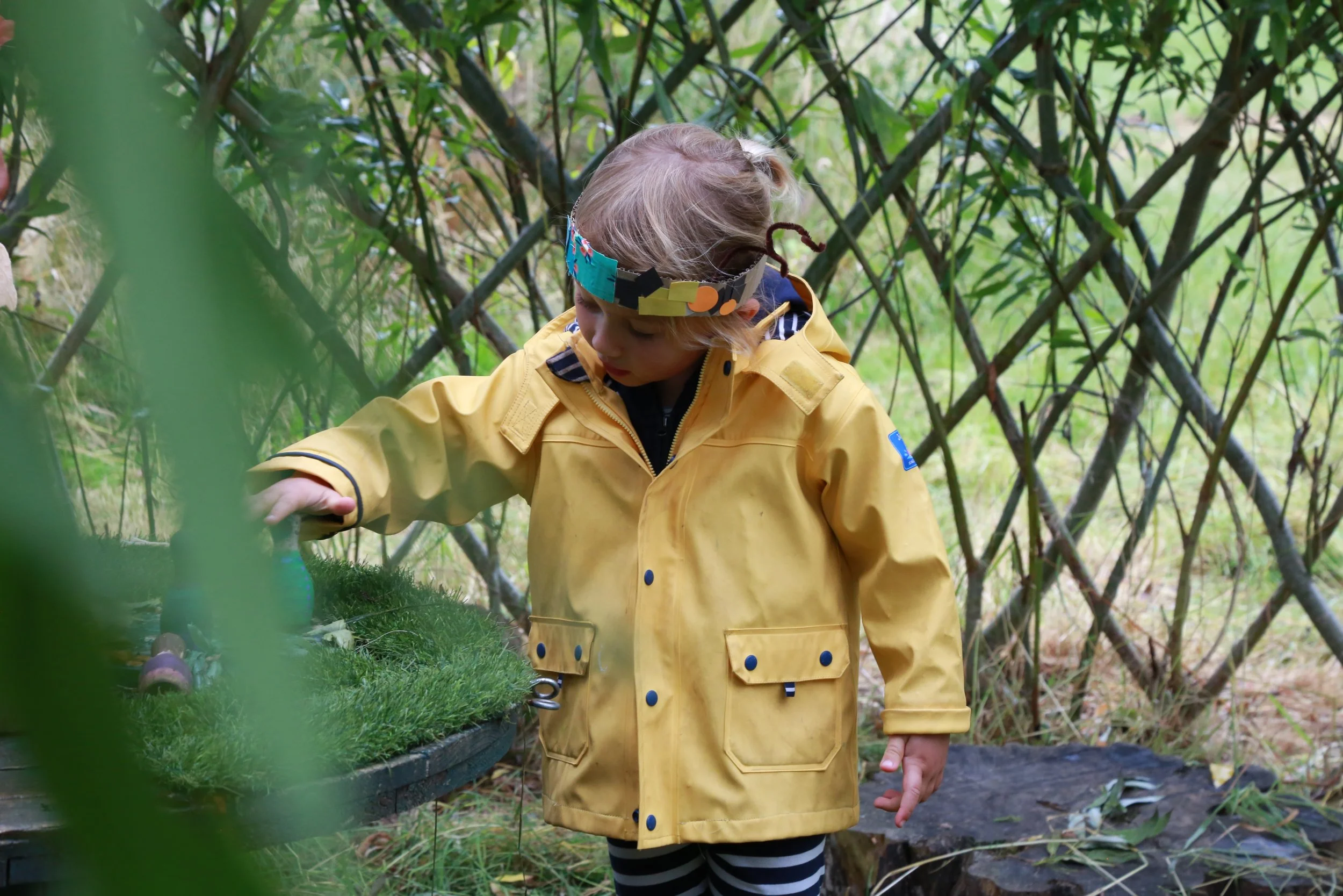 A young girl with blonde hair wearing a colorful handmade paper crown, a yellow raincoat, and striped leggings, exploring outdoors among grass and foliage in a wooded area.