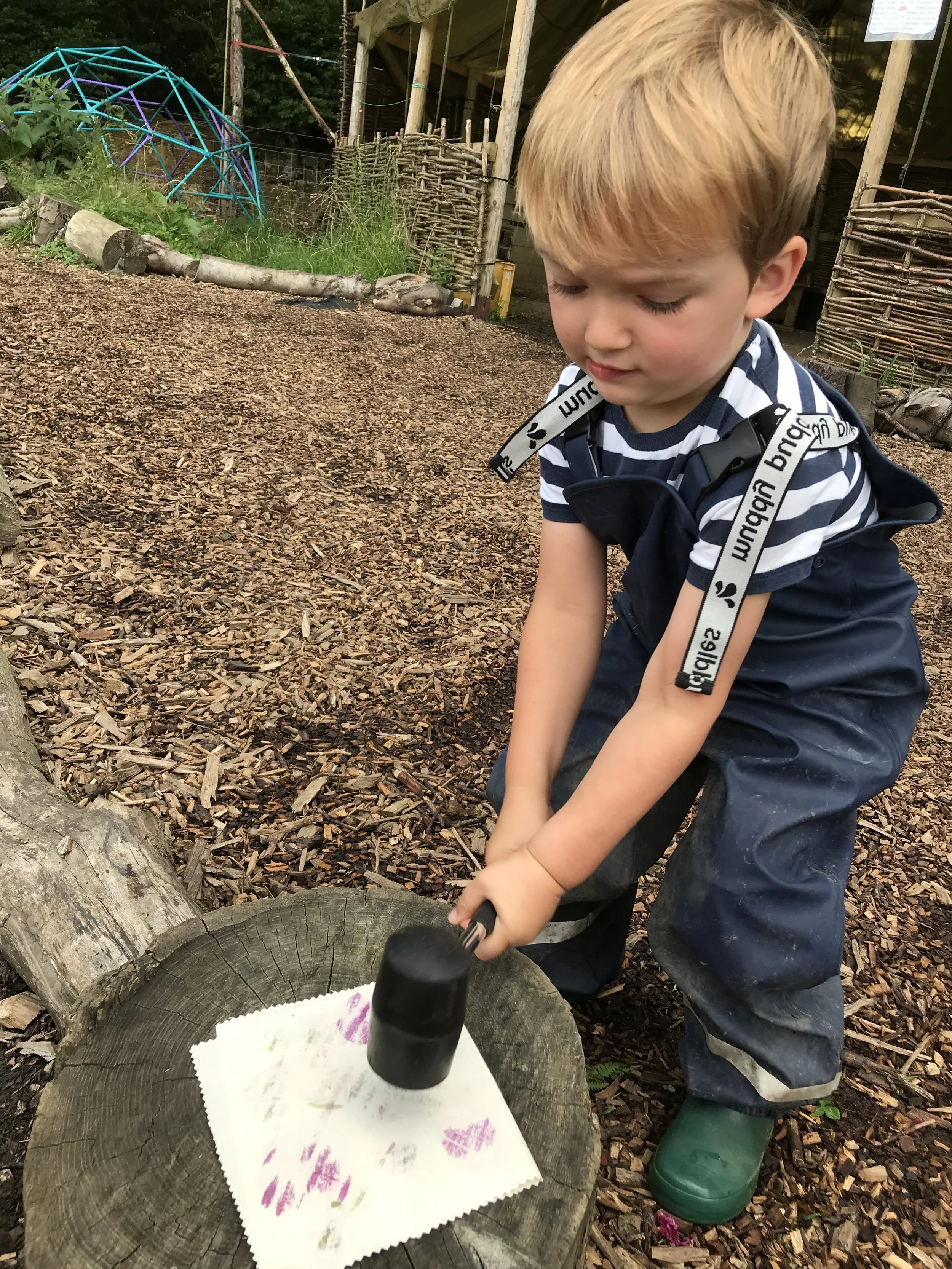 A young boy with blonde hair, wearing a striped shirt and green rain boots, is chopping wood with a mallet. He is indoors at a daycare or preschool, with a wooded outdoor play area in the background, decorated with wood chips and natural materials.